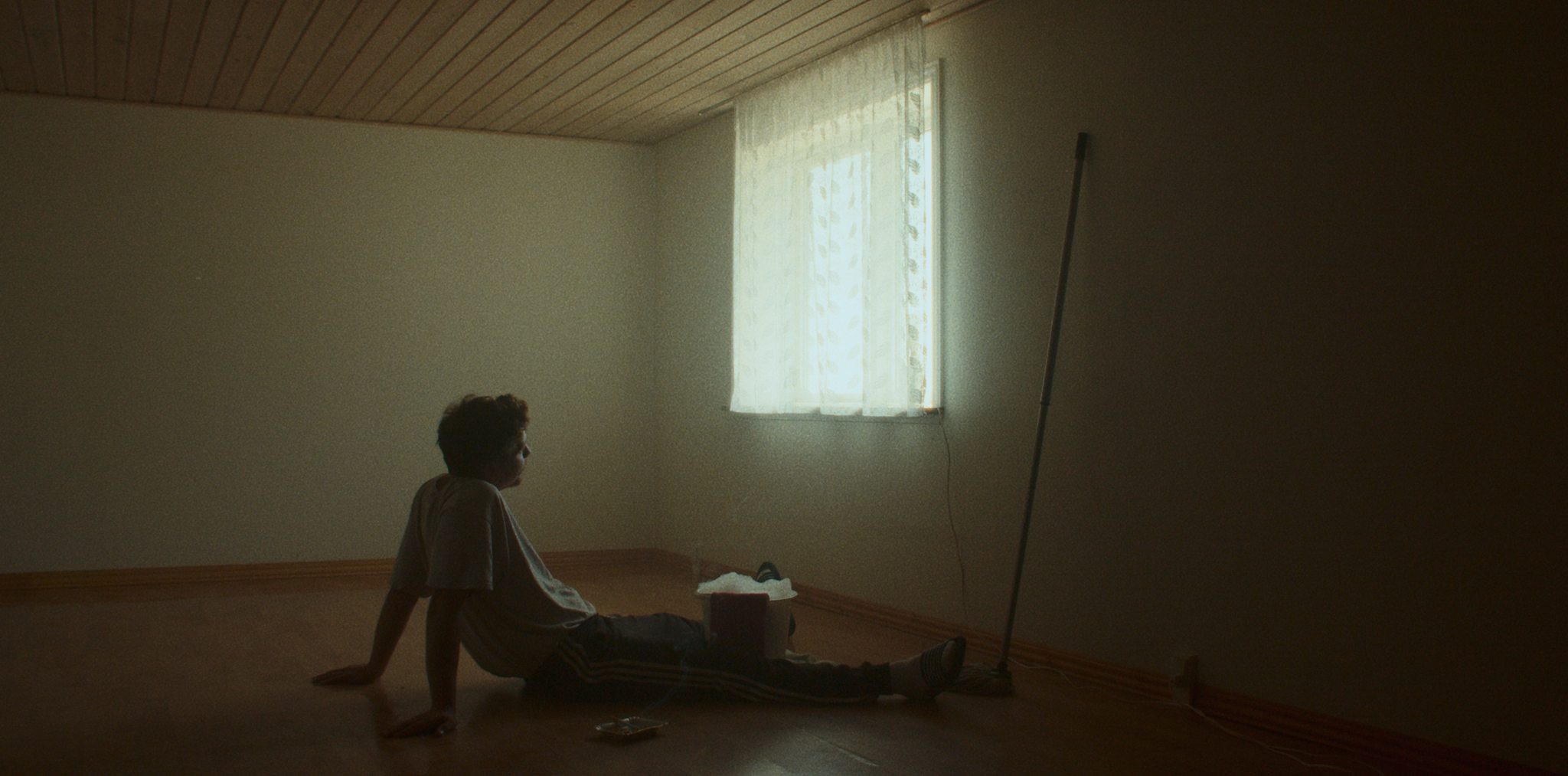 En dreng sidder afslappet på gulvet i et tomt rum med træloft, en enkelt vindue med let gennemskinnelig gardin, mens han kigger ud af vinduet | Kortfilm | Wild Wild Vesløs | Esben Persson | Filmproduk