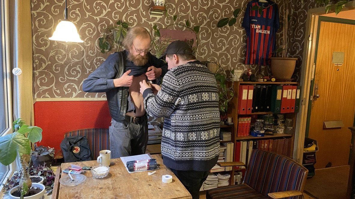 To personer står ved et bord i en stue. Den ene person tager en tatovering af den anden persons mave. Der er bøger og pærer i baggrunden, samt en sports trøje hængende på væggen | Dokumentarfilm | Retrograd | Esben Persson & Anders Mielcke  | Filmpro
