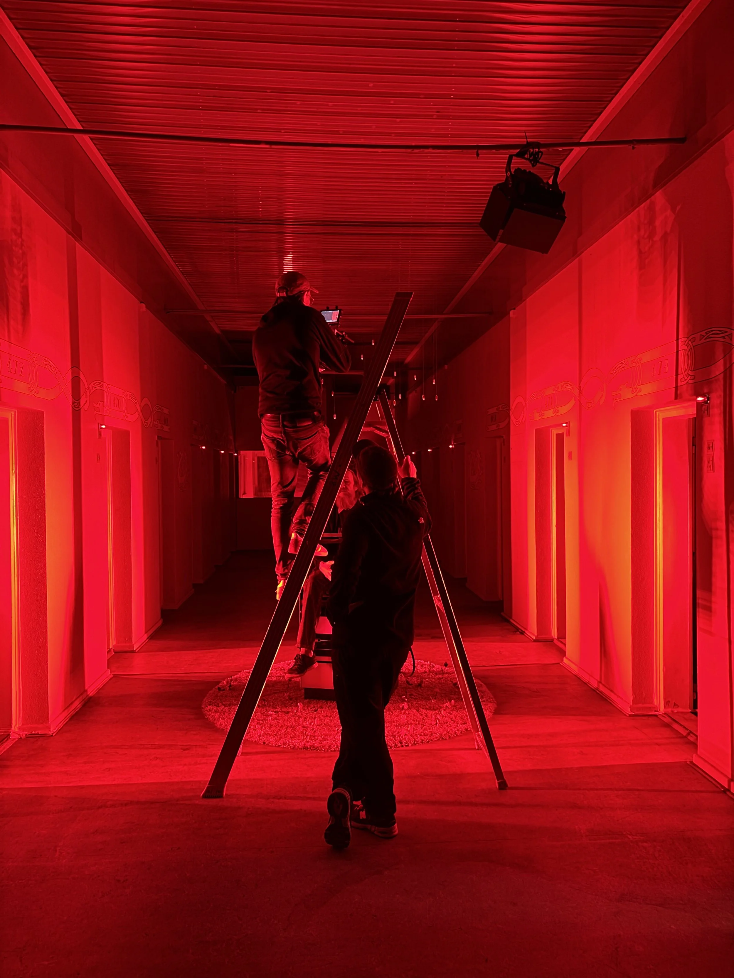 To personer arbejder i en lang, smalt korridor med rød belysning. En person står på en stige og justerer filmlys | Dokumentarfilm | Retrograd | Esben Persson & Anders Mielcke | Filmproduktion Aarhus | GESTALT