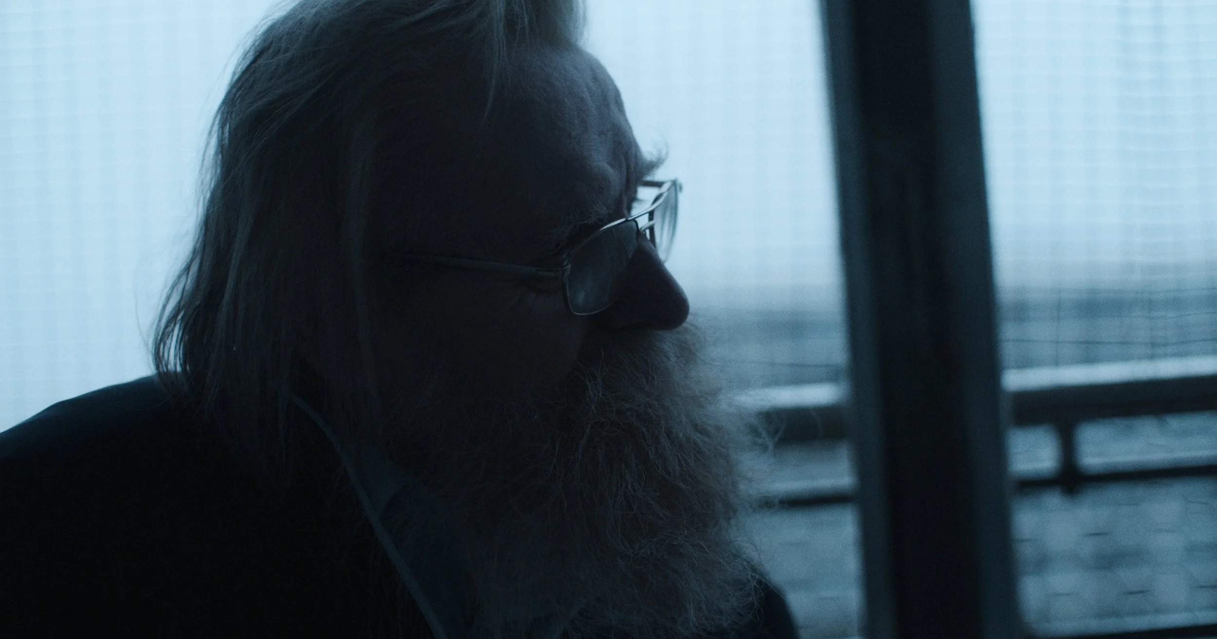 Profilbillede af en ældre mand med langt gråt hår, skæg og briller, der sidder ved et vindue | Dokumentarfilm | Retrograd | Esben Persson & Anders Mielcke  | Filmproduktion Aarhus | GESTALT