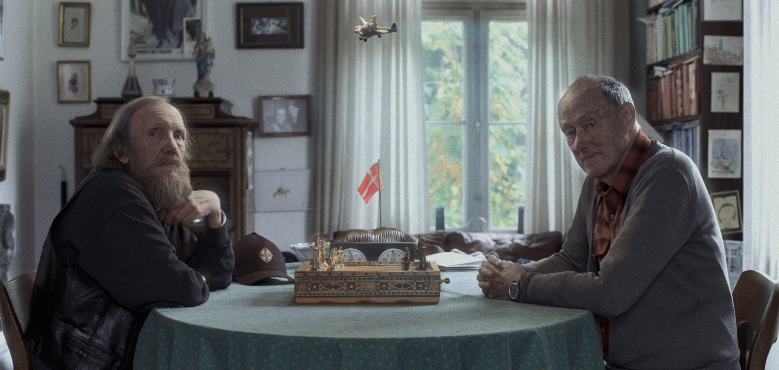 To ældre mænd sidder over for hinanden ved et ovalt bord, der har et skakbræt i midten. Bag dem er der store vinduer med hvide gardiner og bøger på reoler | Dokumentarfilm | Retrograd | Esben Persson & Anders Mielcke | Filmproduktion Aarhus | GESTALT