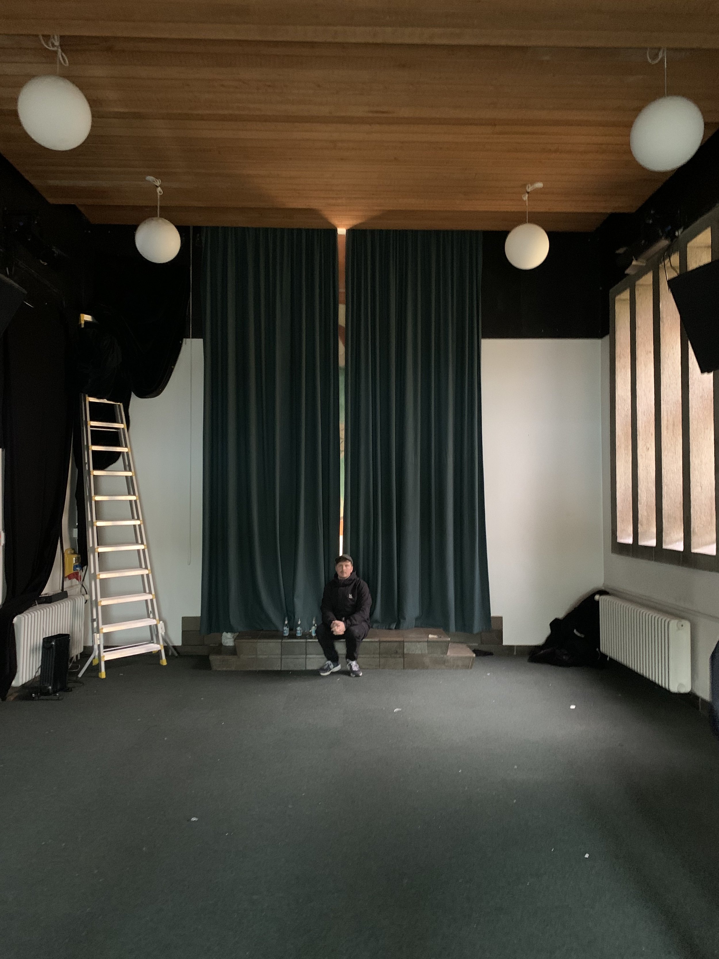 En person sidder foran et stor grønt scenetæppe på en tom indendørs scene med træloft og store vinduer | Dokumentarfilm | Retrograd | Esben Persson & Anders Mielcke | Filmproduktion Aarhus | GESTALT