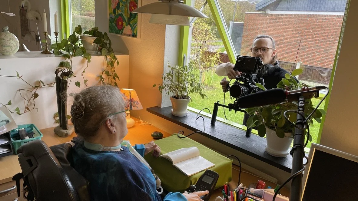 En ældre kvinde sidder ved et skrivebord med en notesbog og et kamera, mens en mand med kamera filmer hende gennem et vindue | Dokumentarfilm | Stemmer fra sær- og åndssvageforsorgen | Anders Mielcke Grønbæk | Filmproduktion Aarhus | GESTALT