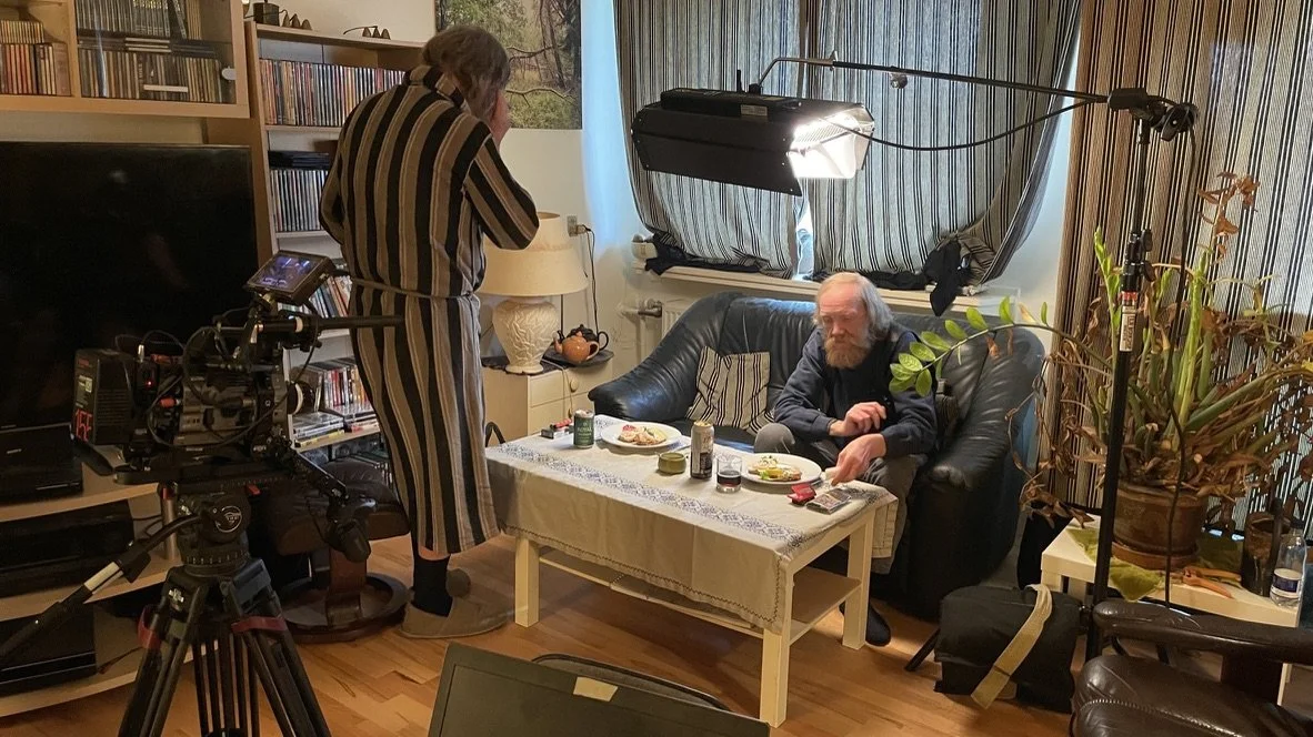 To personer foran et kamera i en stue, hvor den ene sidder på en sofa og spiser, mens den anden står ved siden af. Der er lysudstyr og bøger i baggrunden | Dokumentarfilm | Retrograd | Esben Persson & Anders Mielcke  | Filmproduktion Aarhus | GESTALT