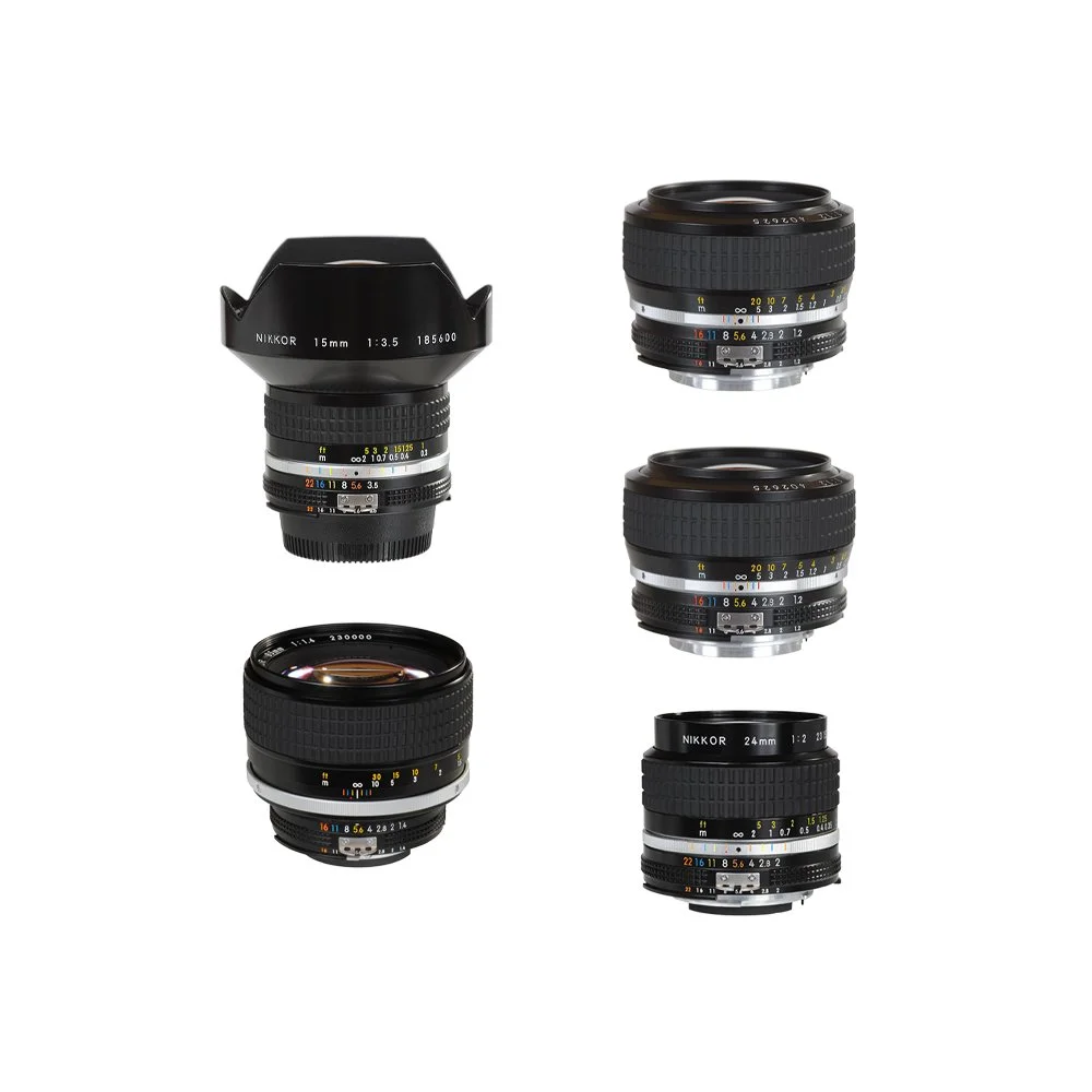 Fem kameraobjektiver, herunder et Nikkor 15mm, og andre optiske linser, placeret på en hvid baggrund.