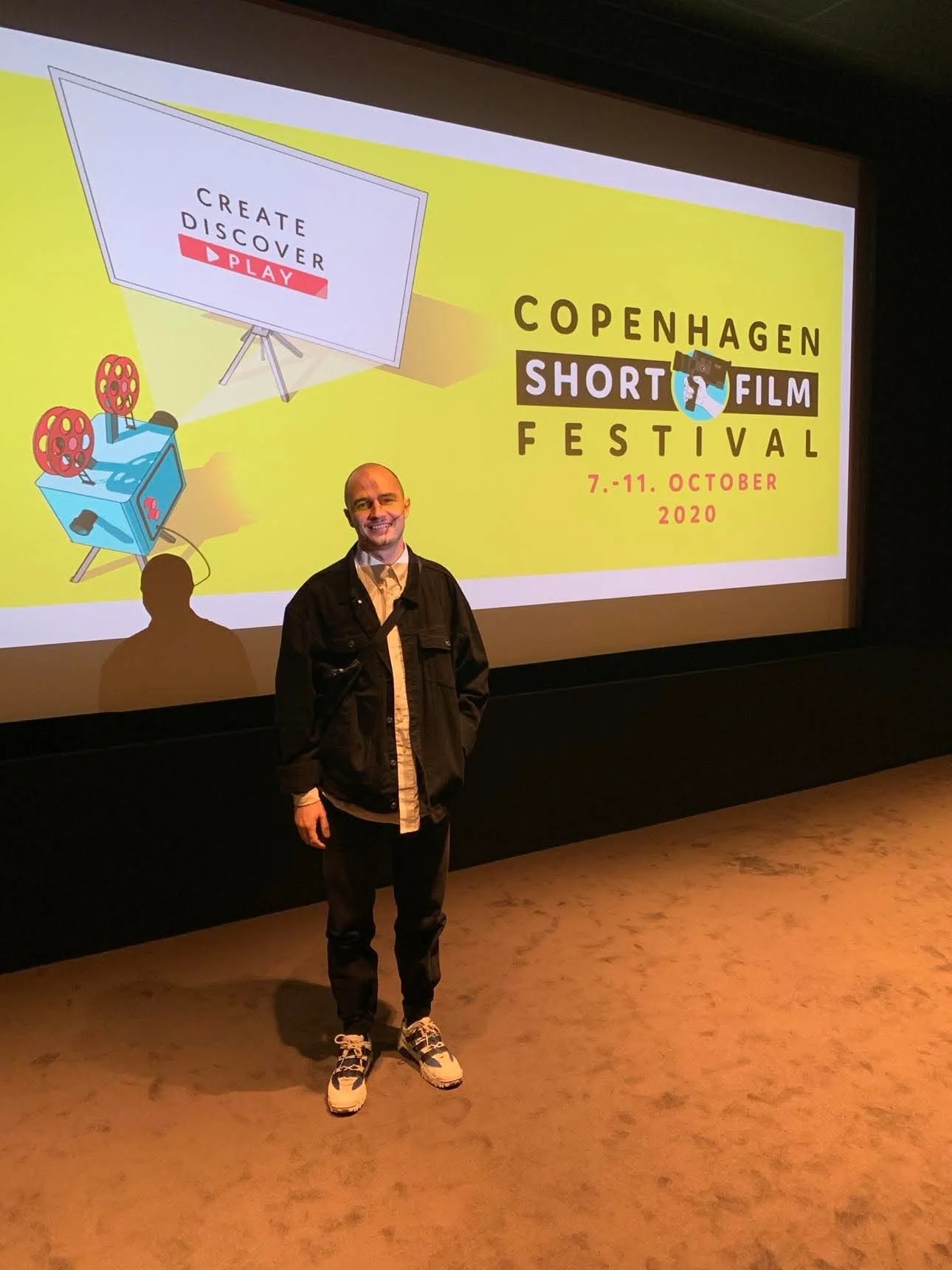 Mand står foran en skærm med plakaten for Københavns Kortfilm Festival 2020, der finder sted 7.-11. oktober, med teksten 'Create, Discover, Play' | Kortfilm | In Medias Ræs | Esben Persson | Filmproduktion Aarhus | GESTALT