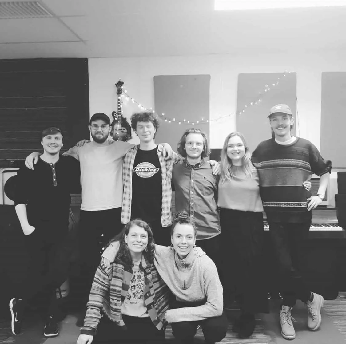 Gruppe af otte mennesker, fem står bag og to er siddende foran, i et musikrummiljø med guitarer og keyboard, alle smilende og har armene om hinanden | Musikkomposition | Studieproduktion og lyddesign | Filmproduktion Aarhus | GESTALT