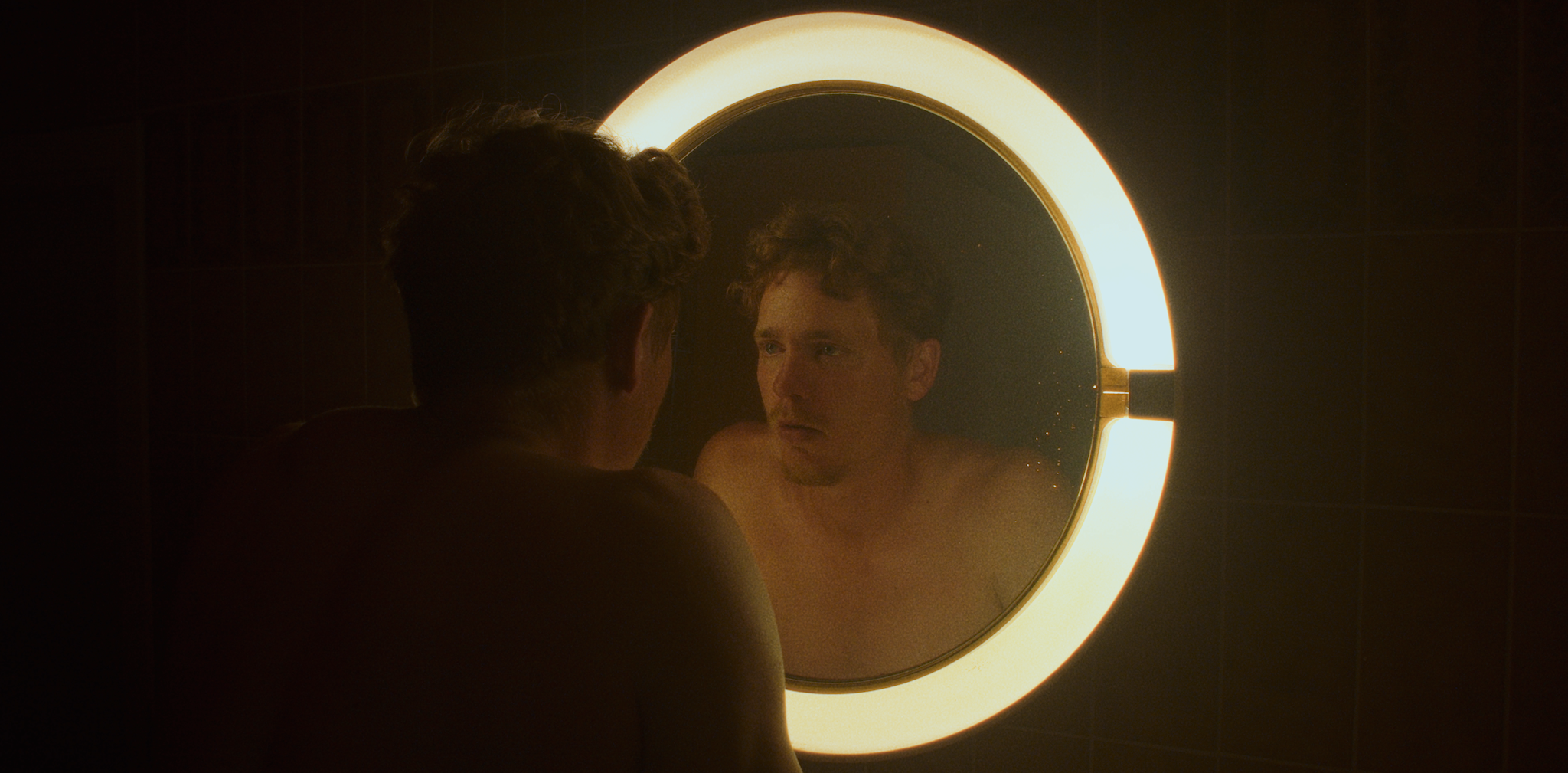En mand ser på sin refleksion i et rundt badeværelses spejl med lys rundt om. Han er uden tøj, og rummet er mørkt med minimal belysning fra spejlets lys | Kortfilm | Wild Wild Vesløs | Esben Persson | Filmproduktion Aarhus | GESTALT