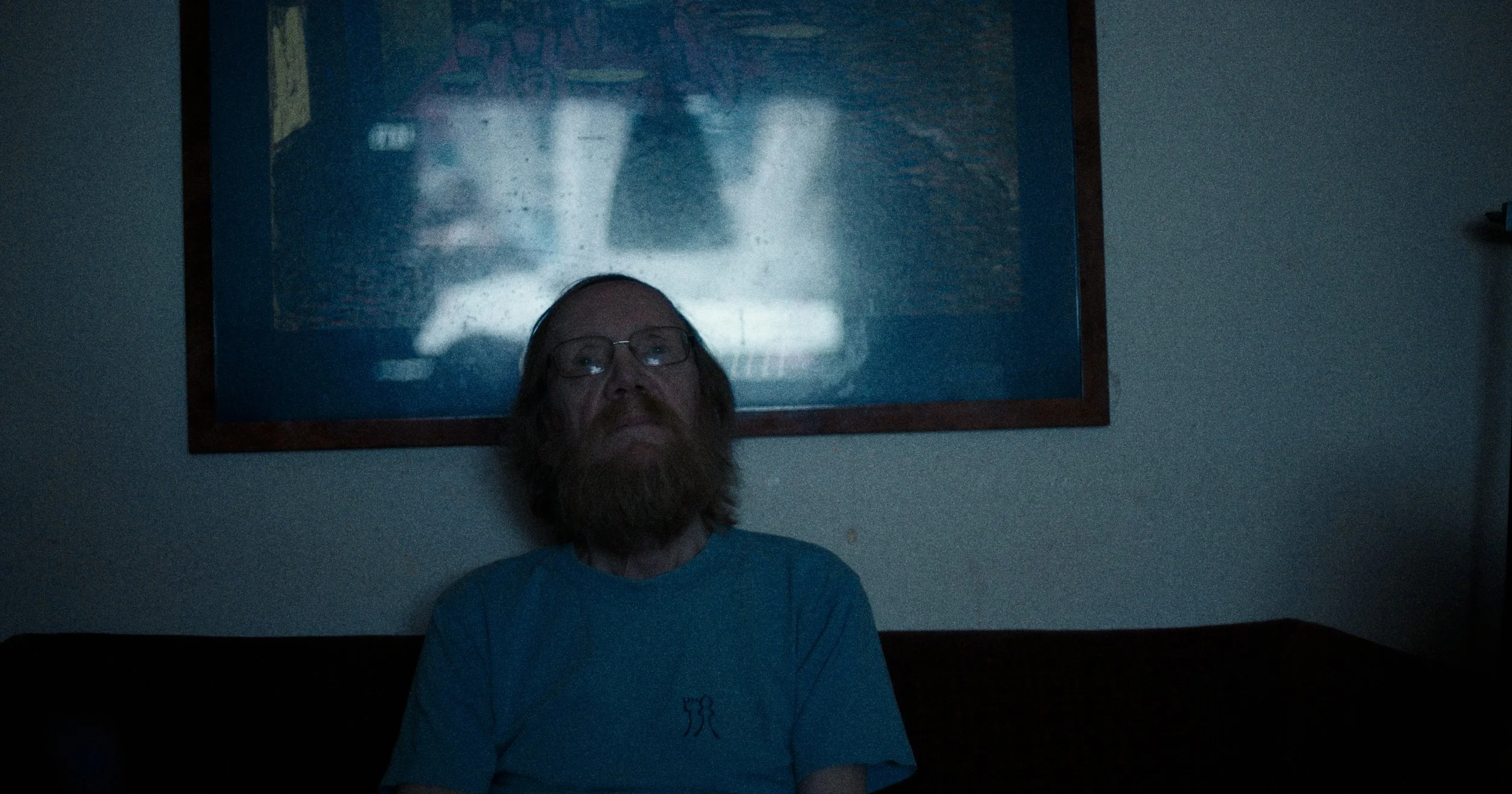 En mand med brunt skæg og briller sidder i et mørkt rum og ser TV | Dokumentarfilm | Retrograd | Esben Persson & Anders Mielcke | Filmproduktion Aarhus | GESTALT