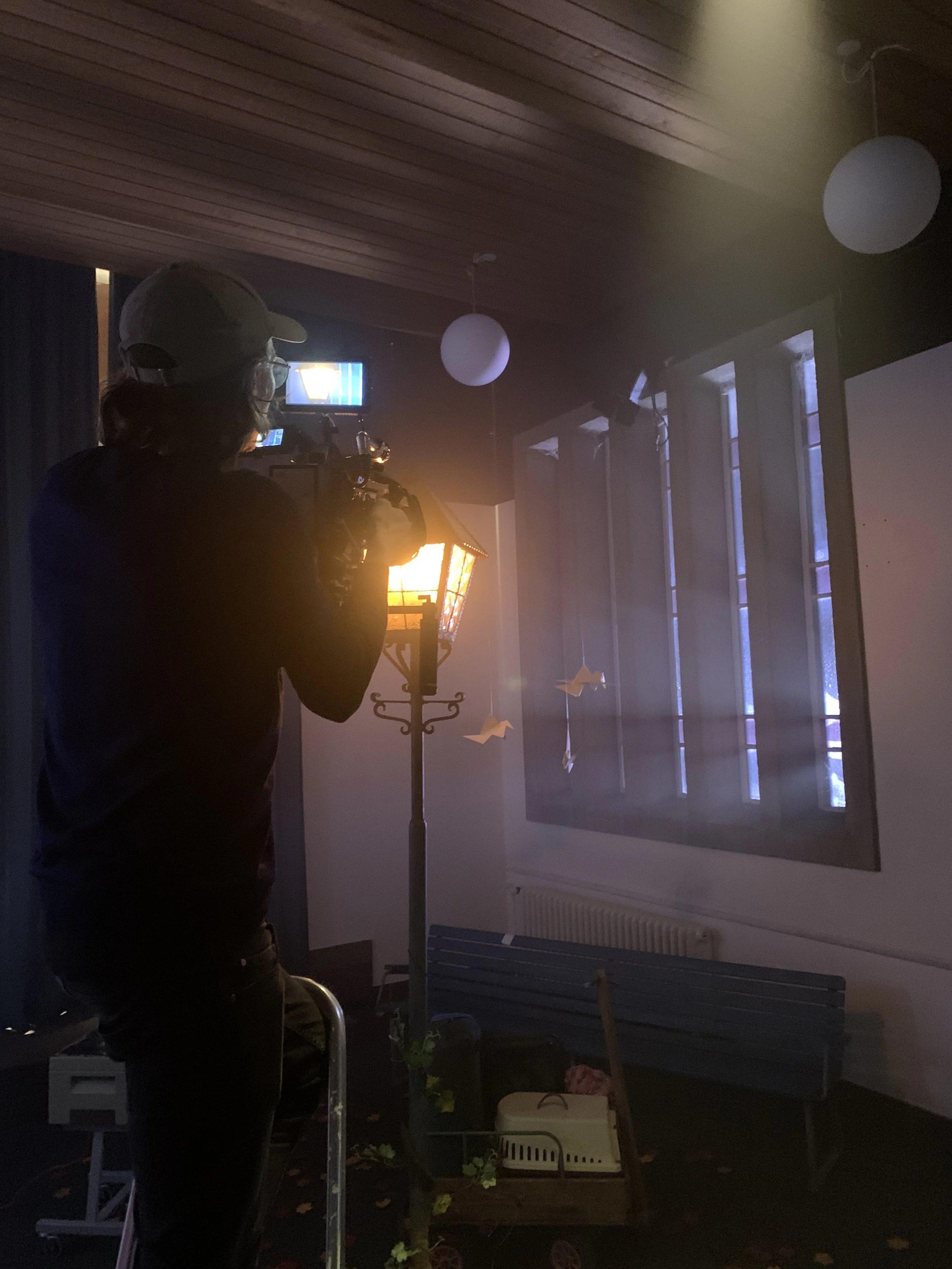 En person med kamera tager billede af en lampe i et mørkt rum med vinduer og papirpynt | Dokumentarfilm | Retrograd | Esben Persson & Anders Mielcke | Filmproduktion Aarhus | GESTALT