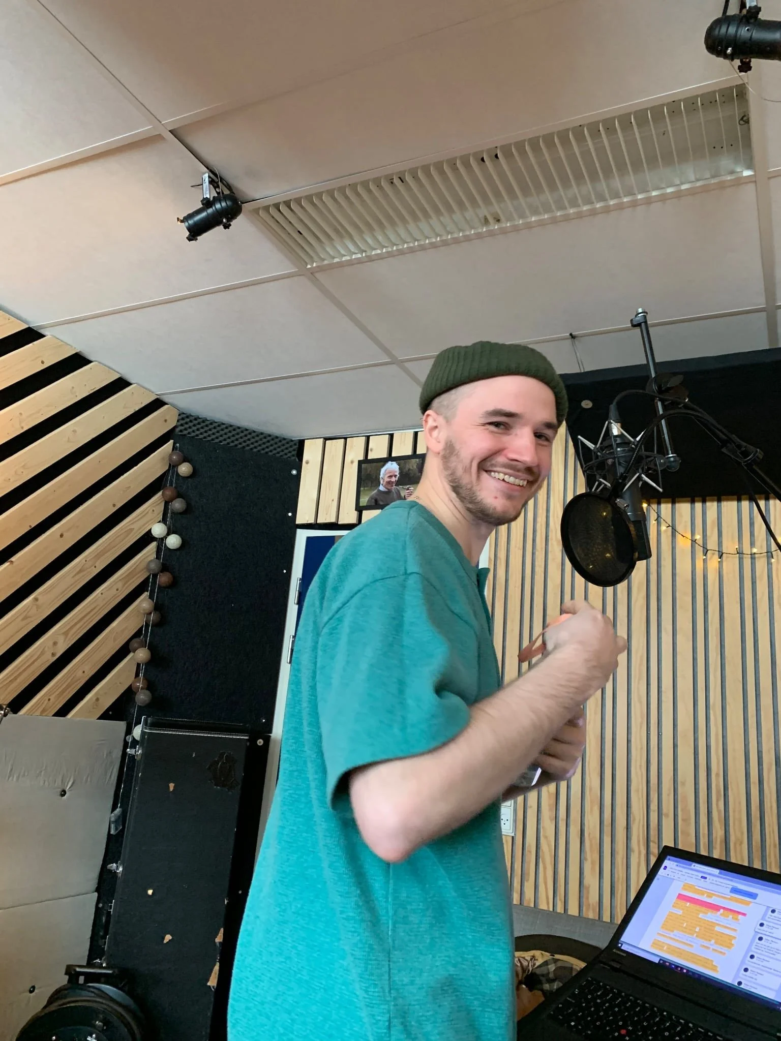 En ung mand med skæg, grøn hue og blå t-shirt står i et lydstudie foran mikrofon, med en åben computer med lydredigeringssoftware på skærmen | Kortfilm | Wild Wild Vesløs | Esben Persson | Filmproduktion Aarhus | GESTALT