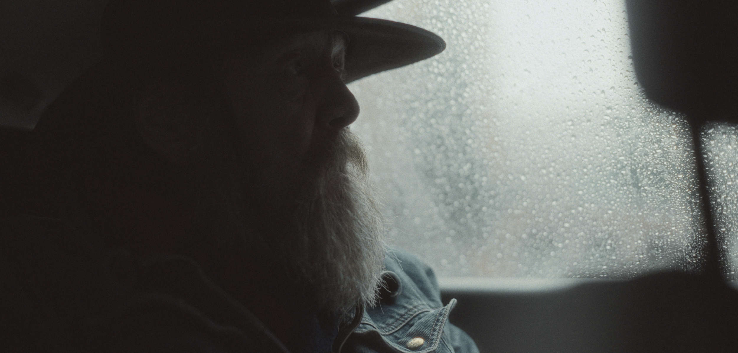 Mand med skæg, hat og jakke sidder i en bil ved regnfuld rude, set i profil | Dokumentarfilm | Retrograd | Esben Persson & Anders Mielcke | Filmproduktion Aarhus | GESTALT