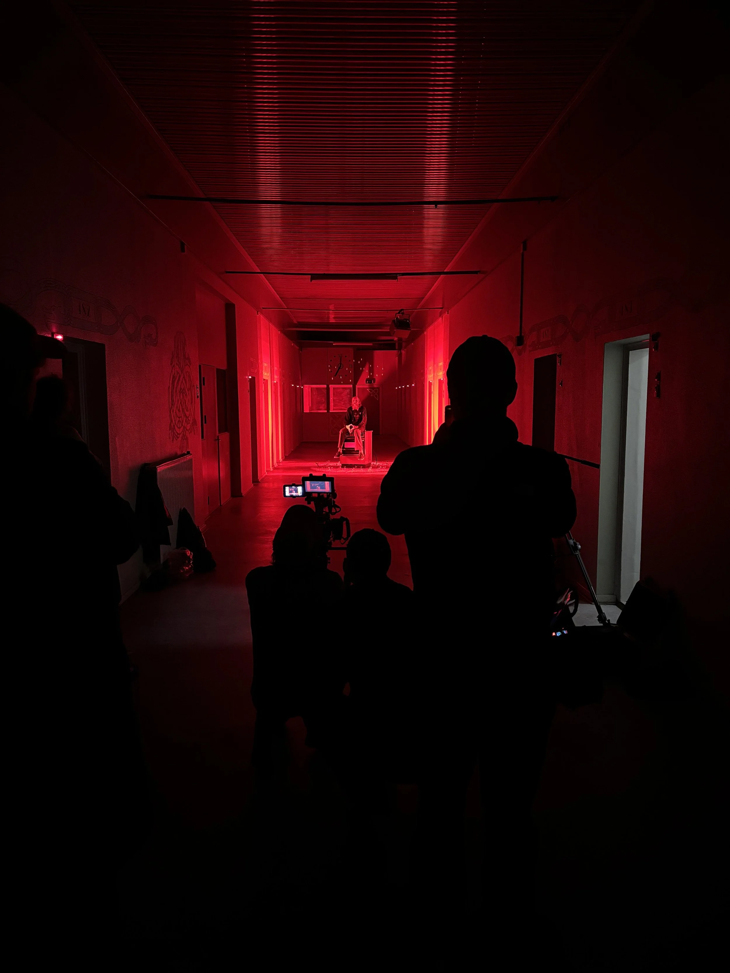 En mørk korridor med rødt lys, hvor en person sidder på en gammel gasovn. Der er filmudstyr, herunder et kamera, rettet mod personen på scenen. | Dokumentarfilm | Retrograd | Esben Persson & Anders Mielcke | Filmproduktion Aarhus | GESTALT