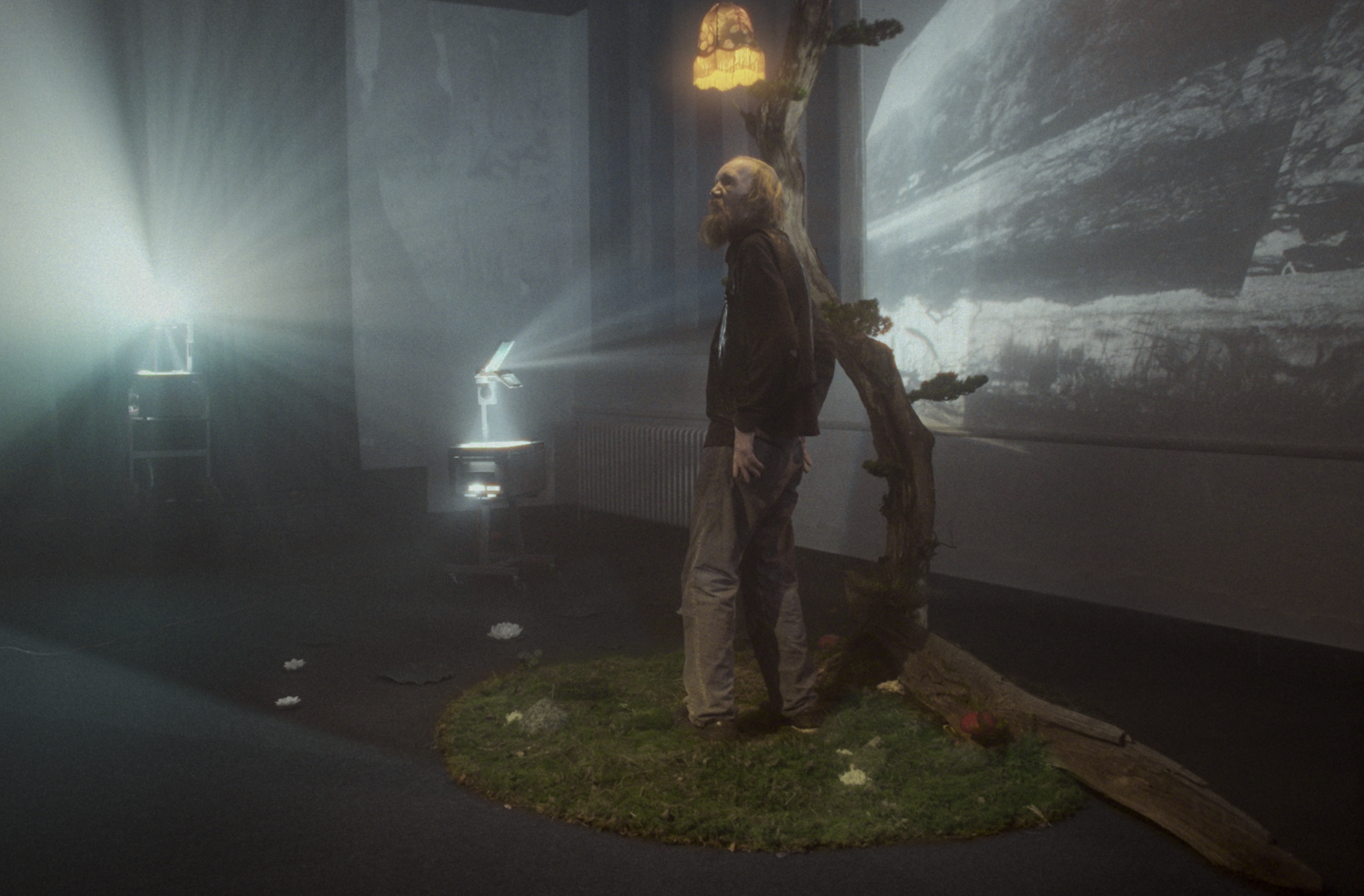 En person med skæg står foran et kunstigt træ i et mørkt filmstudie med projektorer, der viser naturbilleder på væggen | Dokumentarfilm | Retrograd | Esben Persson & Anders Mielcke | Filmproduktion Aarhus | GESTALT