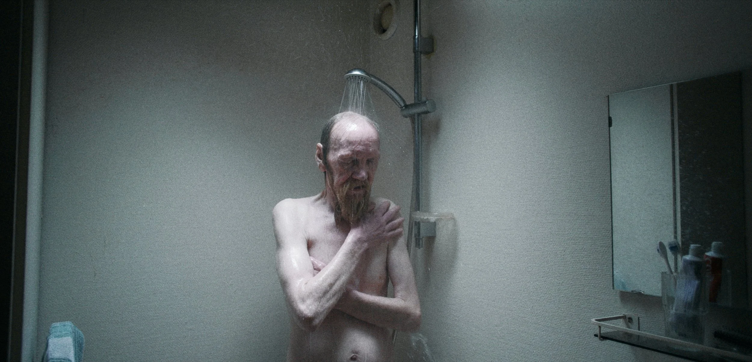 En ældre mand står under bruseren i et badeværelse, med armene korslagt over brystet, og ser ned | Dokumentarfilm | Retrograd | Esben Persson & Anders Mielcke | Filmproduktion Aarhus | GESTALT
