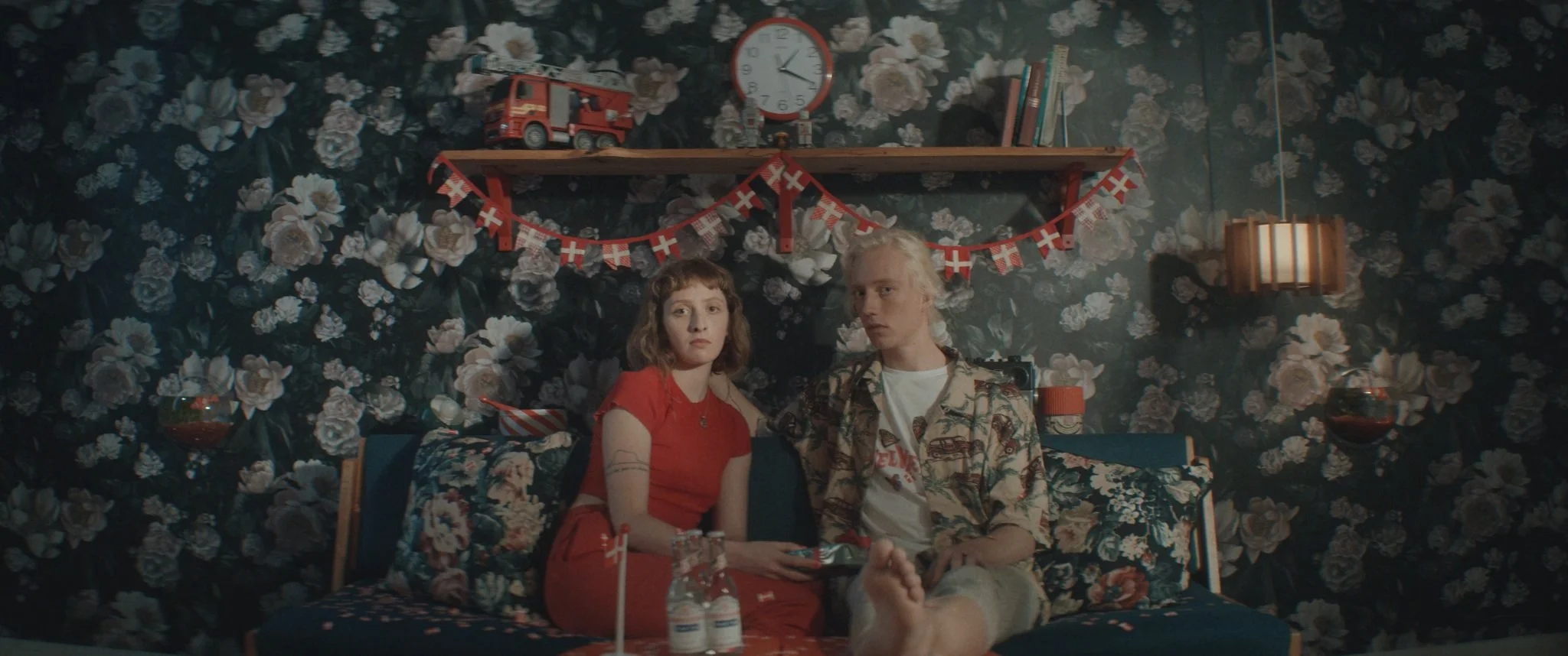 To personer sidder på en seng med blomsterprint, omgivet af festdekorationer såsom små dannebrogflagg, en legemodel af en brandbil, en vægklokke, bøger og væglampe | Kortfilm | In Medias Ræs | Esben Persson | Filmproduktion Aarhus | GESTALT