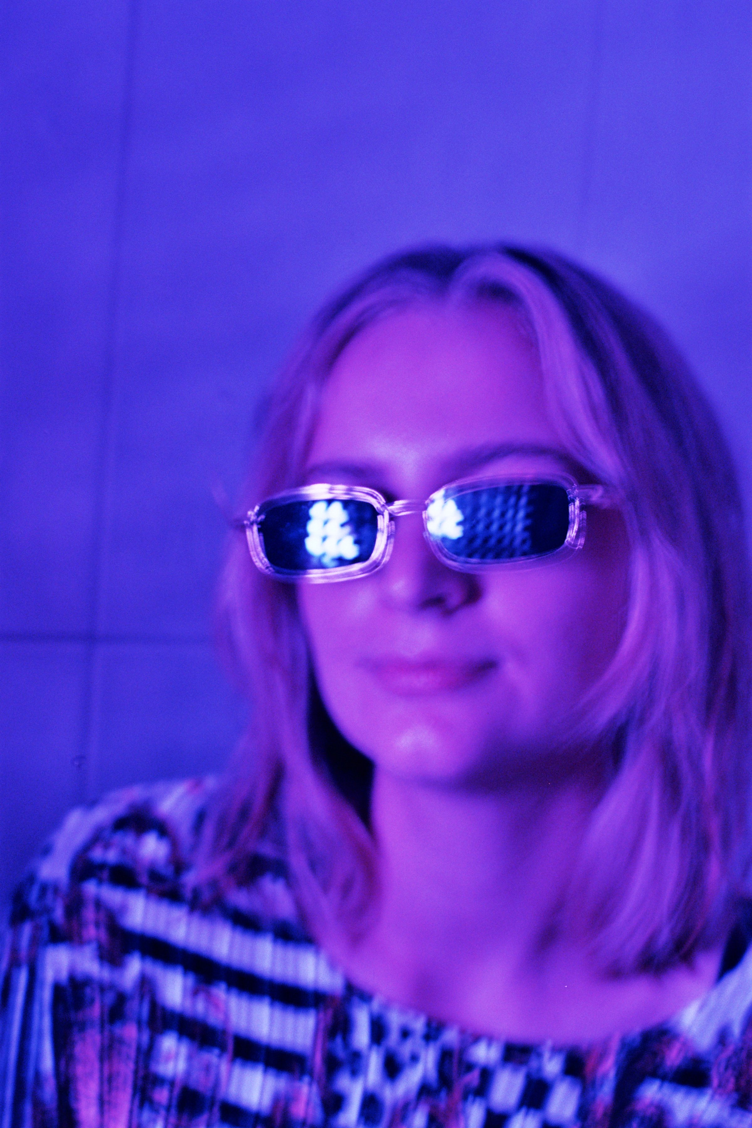 Kvinde med blond hår og solbriller, der reflekterer lyspærer, i en blålilla oplyst baggrund | Location scouting Berlin | Kodak | Filmproduktion Aarhus | GESTALT