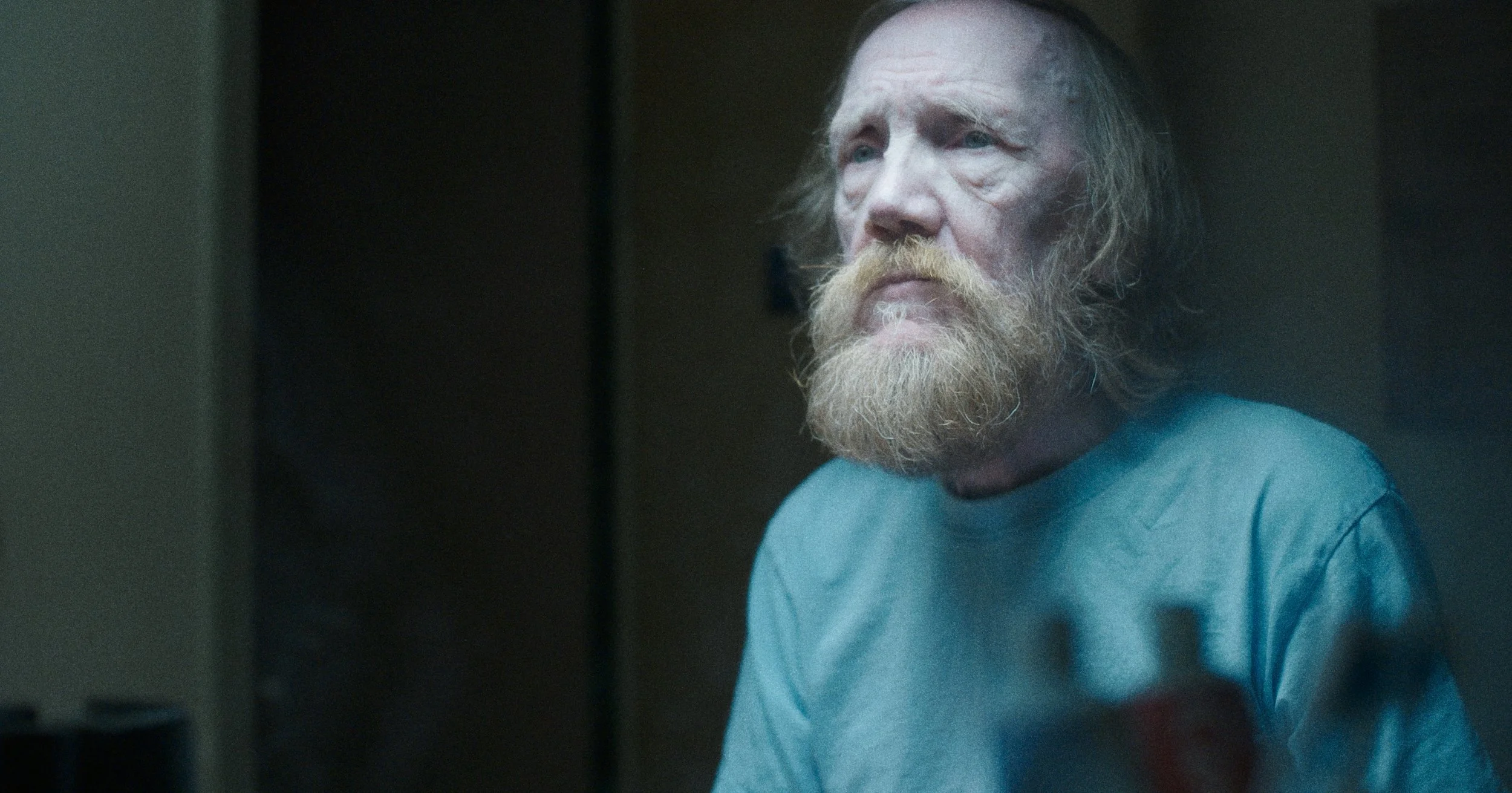 En gammel mand med rødbrunt hår og skæg, der ser trist ud, står i et mørkt rum i en blå T-shirt | Dokumentarfilm | Retrograd | Esben Persson & Anders Mielcke | Filmproduktion Aarhus | GESTALT