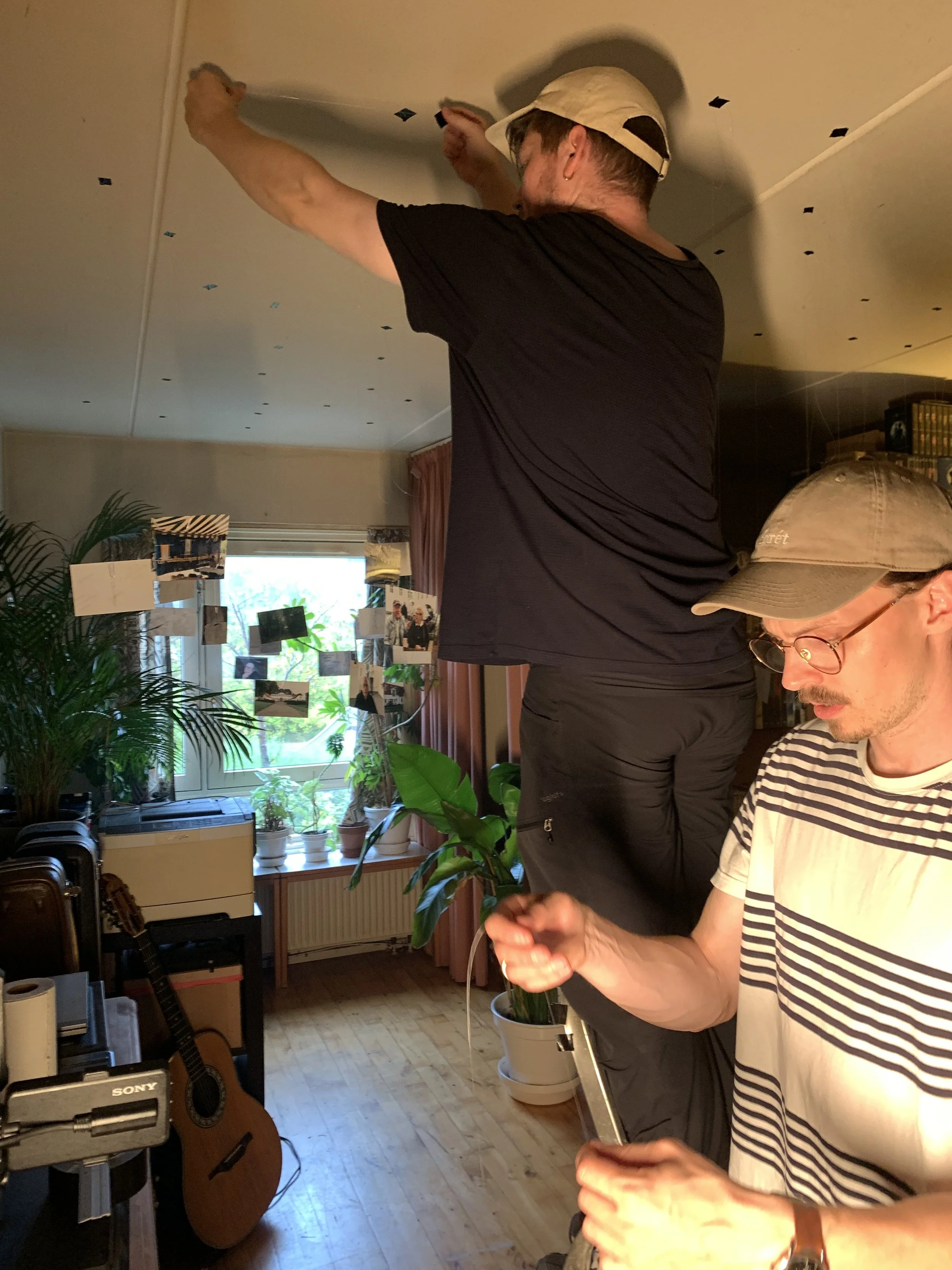 To personer arbejder på at installere et scenerum. Der er planter og fotografier i baggrunden | Dokumentarfilm | Retrograd | Esben Persson & Anders Mielcke | Filmproduktion Aarhus | GESTALT