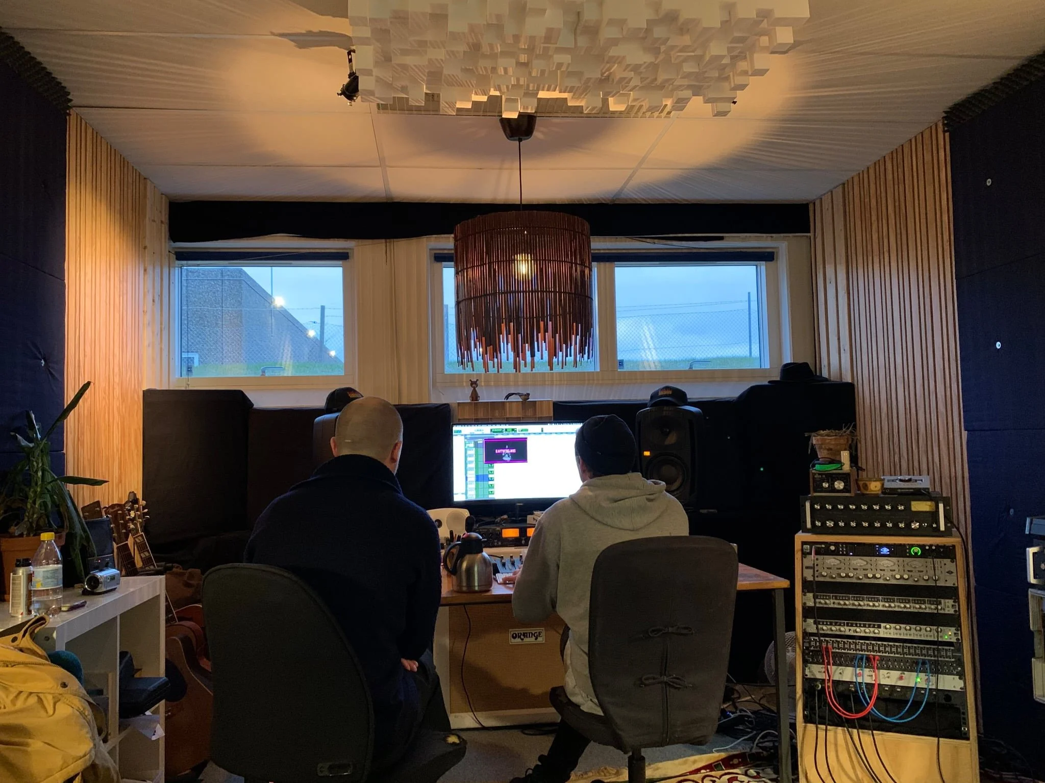 To personer arbejder i et lydstudie, foran en computer med lydudstyr, omgivet af lydudstyr og instrumenter. Der er træpaneler på væggene og store vinduer bagved | Musikkomposition | Studieproduktion og lyddesign | Filmproduktion Aarhus | GESTALT