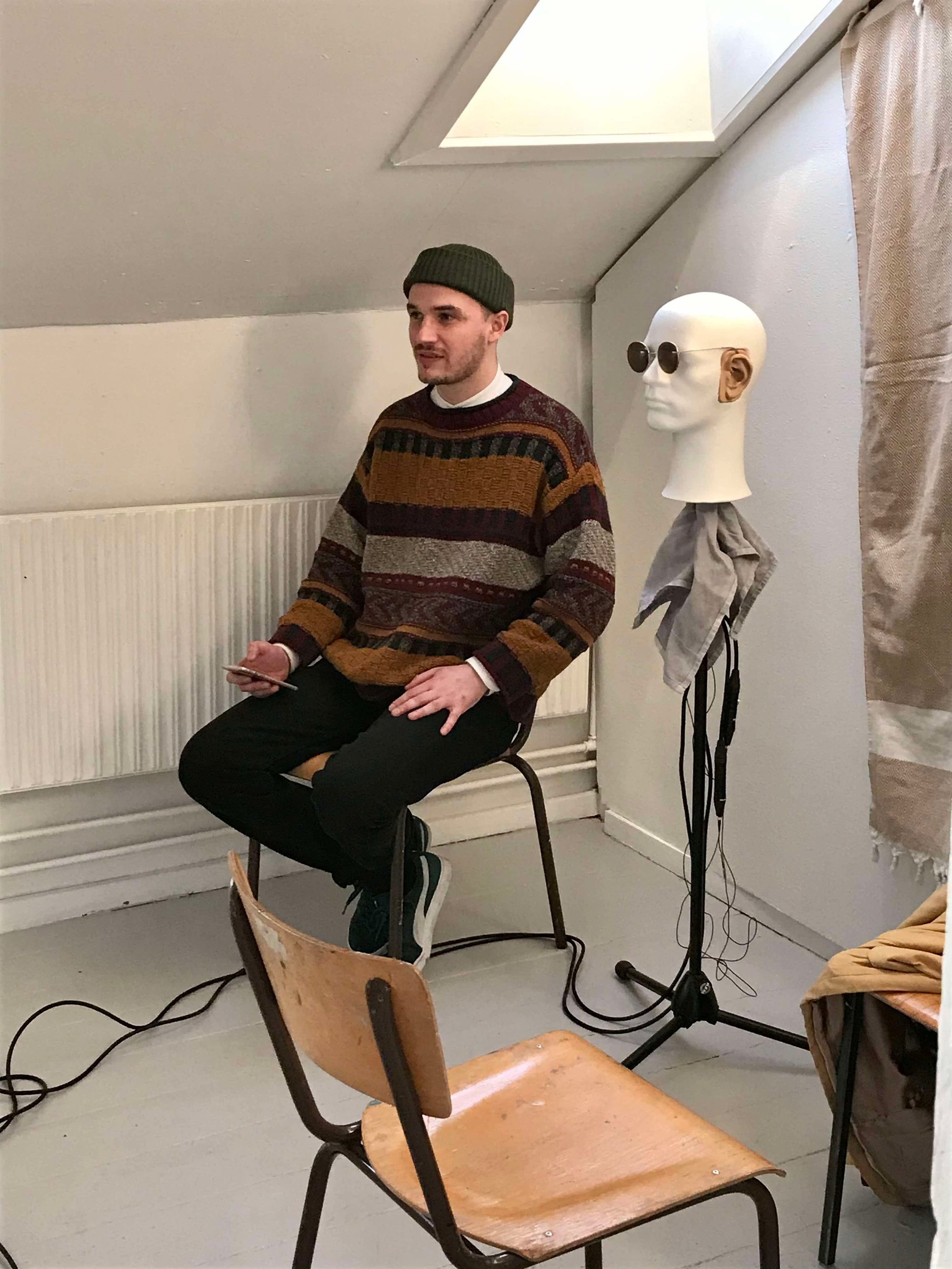En mand sidder på en træstol i et rum med en mannequin hoved, der har solbriller og høretelefoner | Musikkomposition | Studieproduktion og lyddesign | Filmproduktion Aarhus | GESTALT