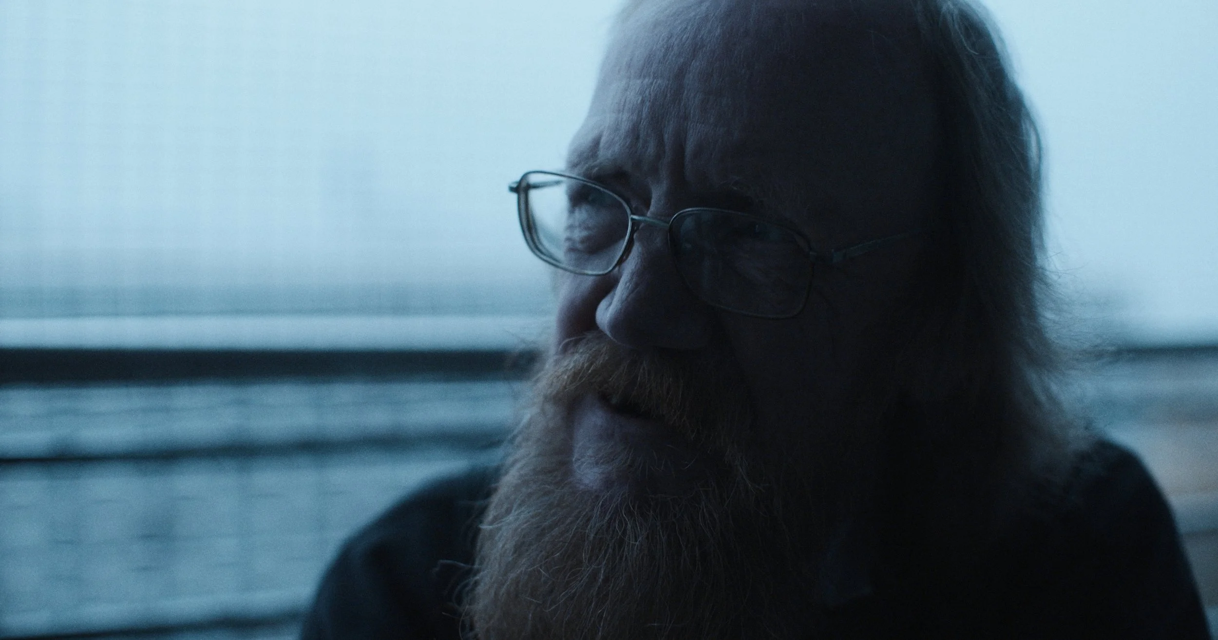 Et portræt af en ældre mand med briller og fuldskæg, set i profil mod en udendørs baggrund. Manden har gråt hår og rødbrunt skæg | Dokumentarfilm | Retrograd | Esben Persson & Anders Mielcke | Filmproduktion Aarhus | GESTALT