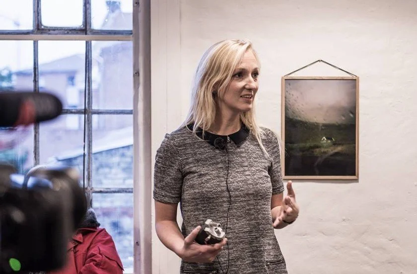 Lærke Posselt taler, mens hun holder en mikrofon. Hu står indendørs foran et hvidt væg med et landskabsbillede  | Film- og fotofestival på Aarhus Kunstakademi ved navn Aarhus Independent Pixels | Eventbilleder | Filmproduktion Aarhus | GESTALT