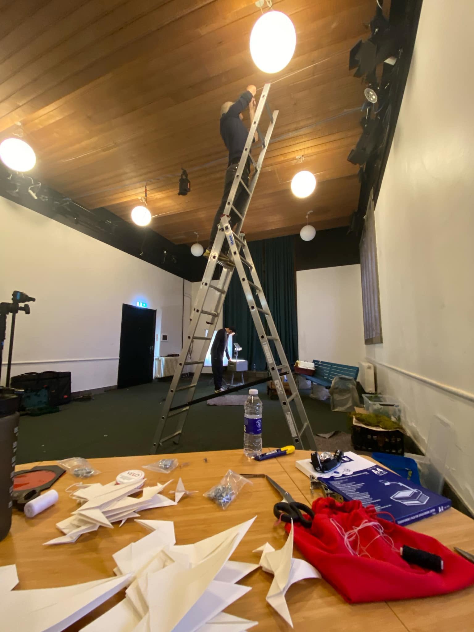 To personer arbejder i et studie, hvor en står på en stige og arbejder i loftet. Der er værktøj og materialer spredt ud over et bord i forgrunden. | Dokumentarfilm | Retrograd | Esben Persson & Anders Mielcke | Filmproduktion Aarhus | GESTALT