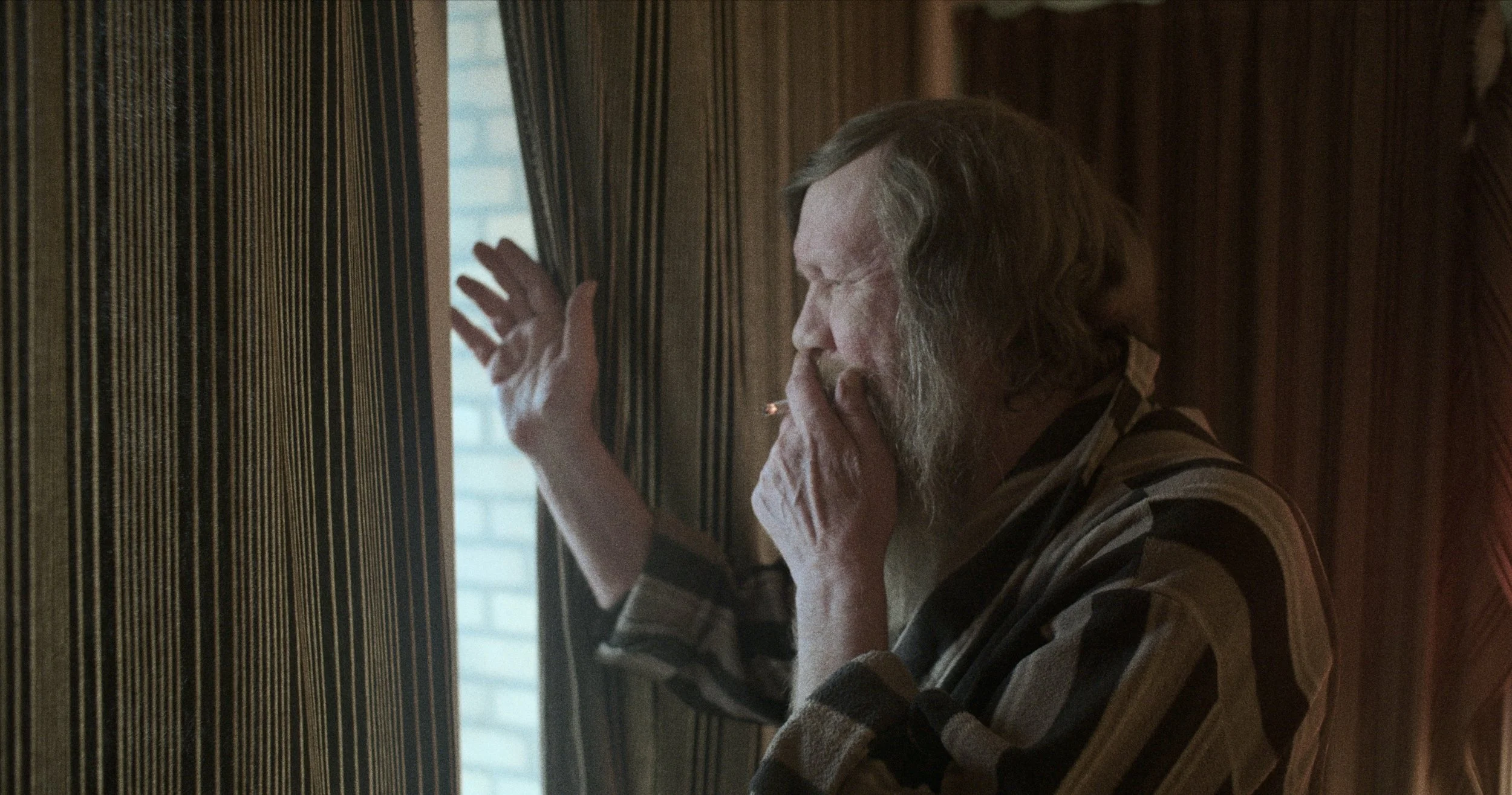 En ældre mand med lang, grå hår og skæg, der ryger en cigaret, mens han kigger ud af et vindue, med en mørk og retro indretning | Dokumentarfilm | Retrograd | Esben Persson & Anders Mielcke  | Filmproduktion Aarhus | GESTALT