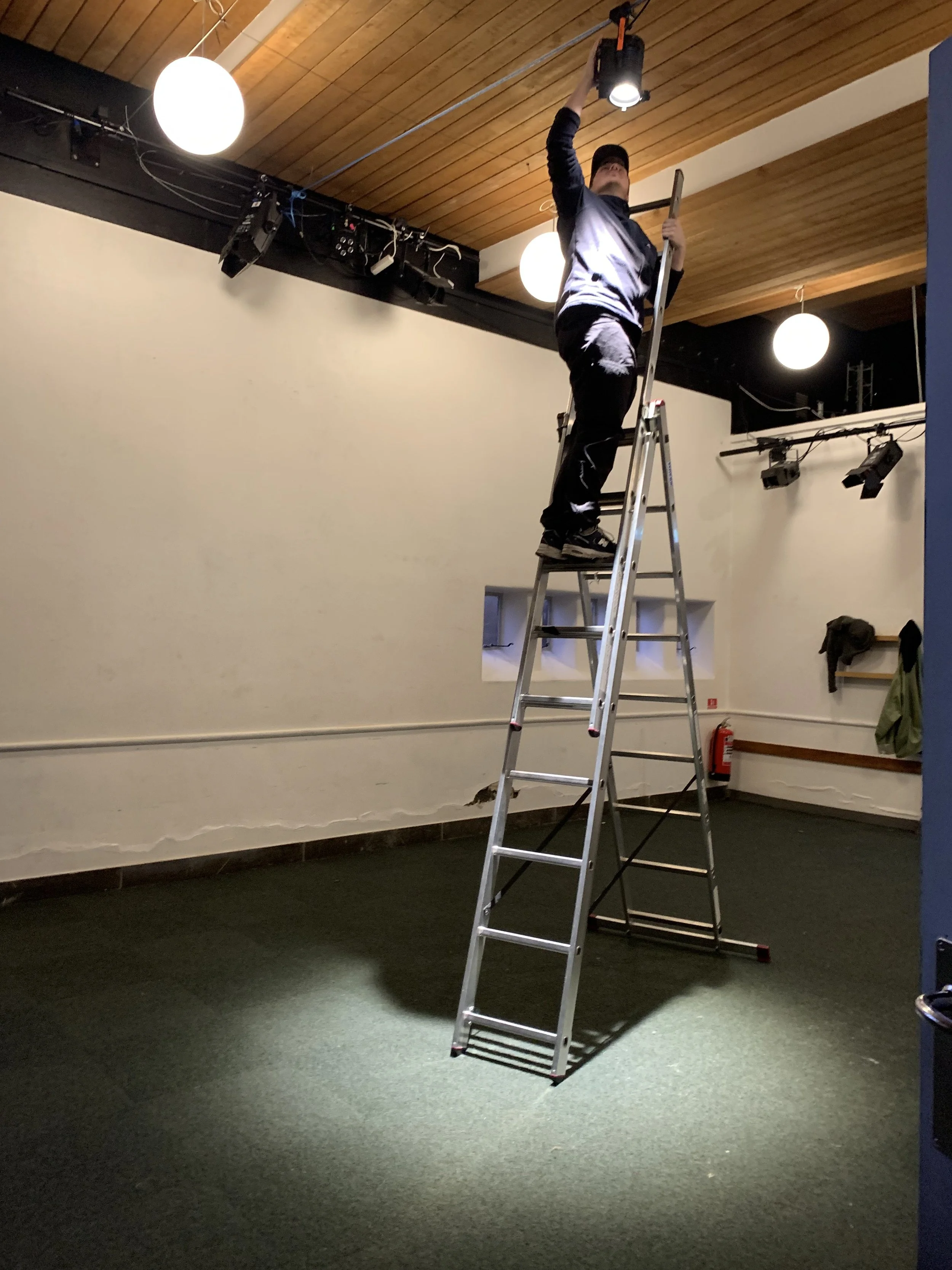 Person står på en stige, og arbejder med en lampe i loftet. Indendørs, med træloft og mørkt gulvtæppe | Dokumentarfilm | Retrograd | Esben Persson & Anders Mielcke | Filmproduktion Aarhus | GESTALT