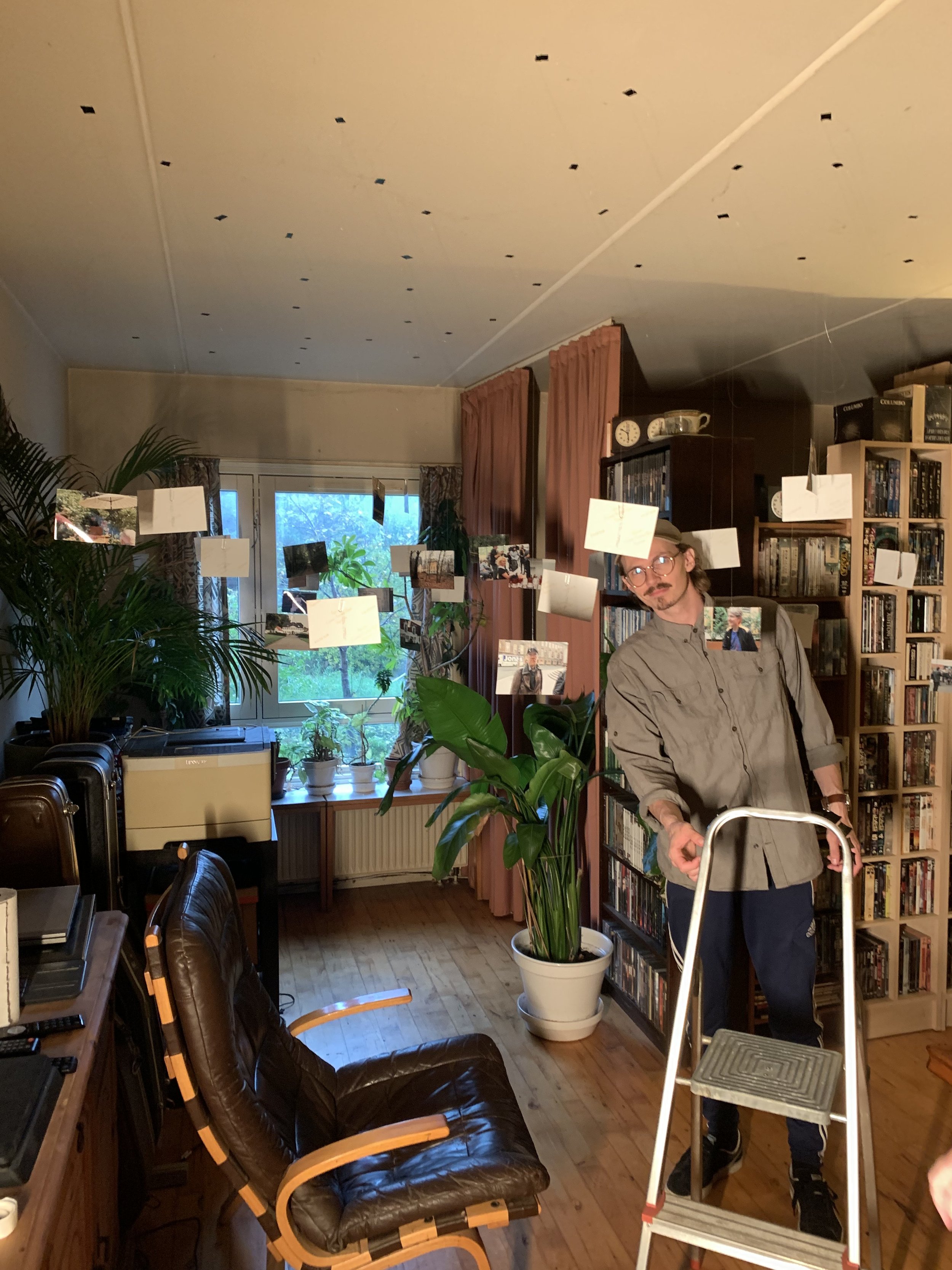 Et indendørs rum med planter, bøger og fotografier. En person står ved en stige, omgivet af pynt og møbler | Dokumentarfilm | Retrograd | Esben Persson & Anders Mielcke | Filmproduktion Aarhus | GESTALT