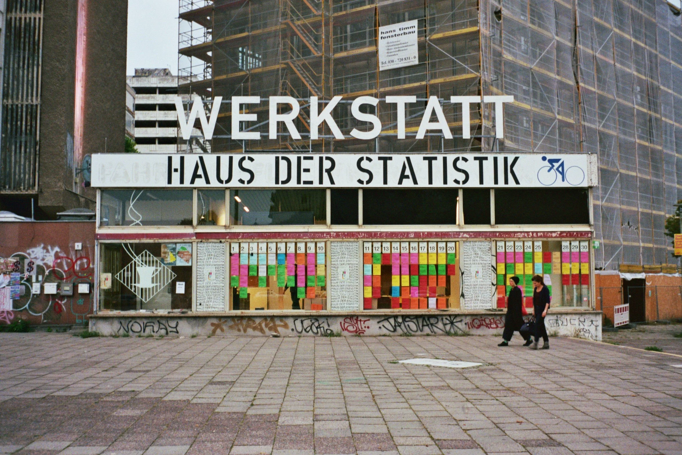 En bygning med store skilte, hvor der står 'Werkstatt' og 'Haus der Statistik'. I fronten er der en væg med farverige notatark og graffitidekorer. To kvinder går forbi på fortovet | Location scouting Berlin | Kodak | Filmproduktion Aarhus | GESTALT