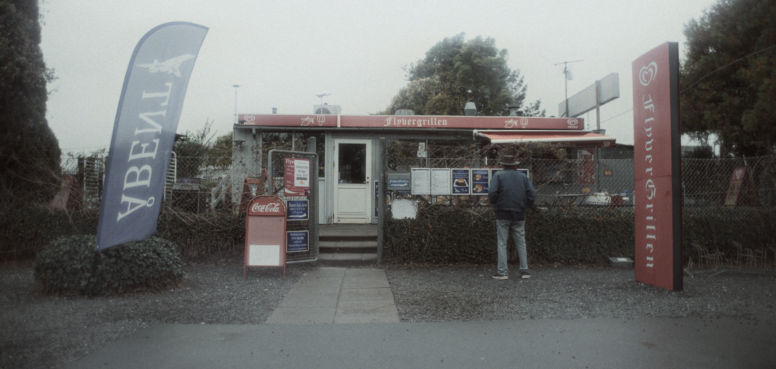 En ensom fodgænger står foran en lille grill med skiltet 'Flyvergrillen' udenfor, omgivet af flag og plakater | Dokumentarfilm | Retrograd | Esben Persson & Anders Mielcke | Filmproduktion Aarhus | GESTALT