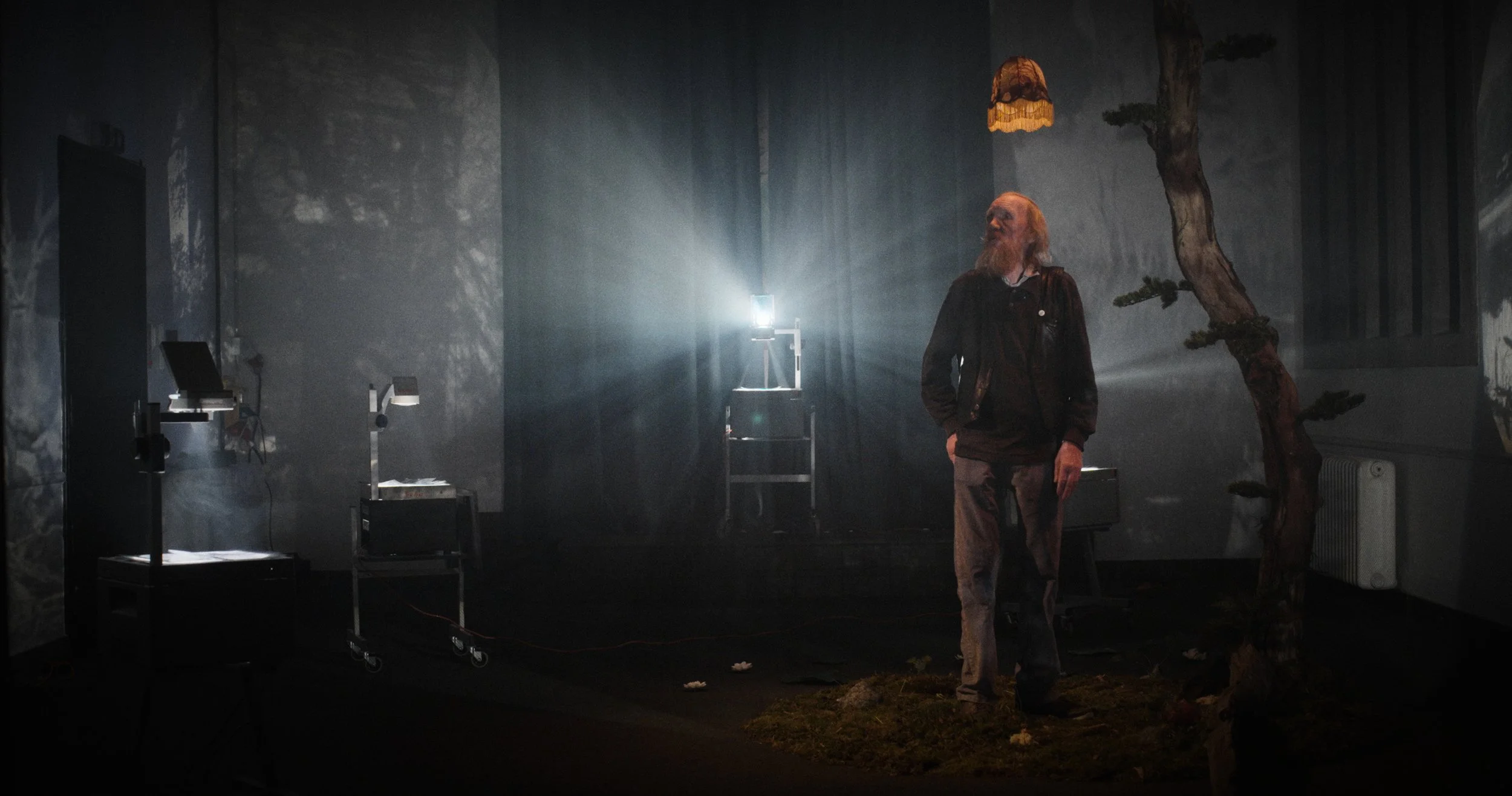 En mand med skæg står i et mørkt rum med kunstig trædekor og en projektor | Dokumentarfilm | Retrograd | Esben Persson & Anders Mielcke | Filmproduktion Aarhus | GESTALT