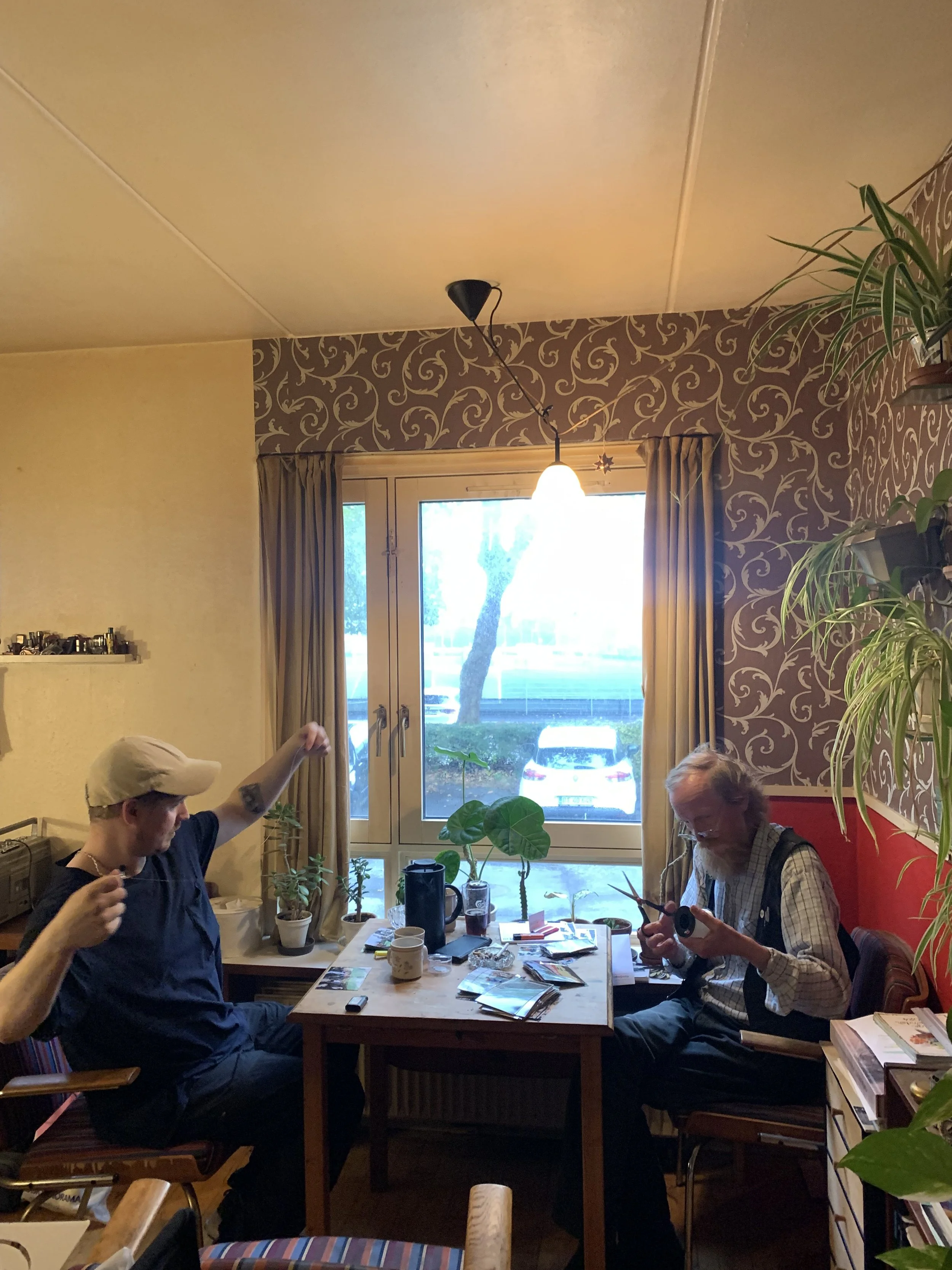 To mænd sidder ved et bord i en stue med planter, bøger og dagligdagsgenstande | Dokumentarfilm | Retrograd | Esben Persson & Anders Mielcke | Filmproduktion Aarhus | GESTALT