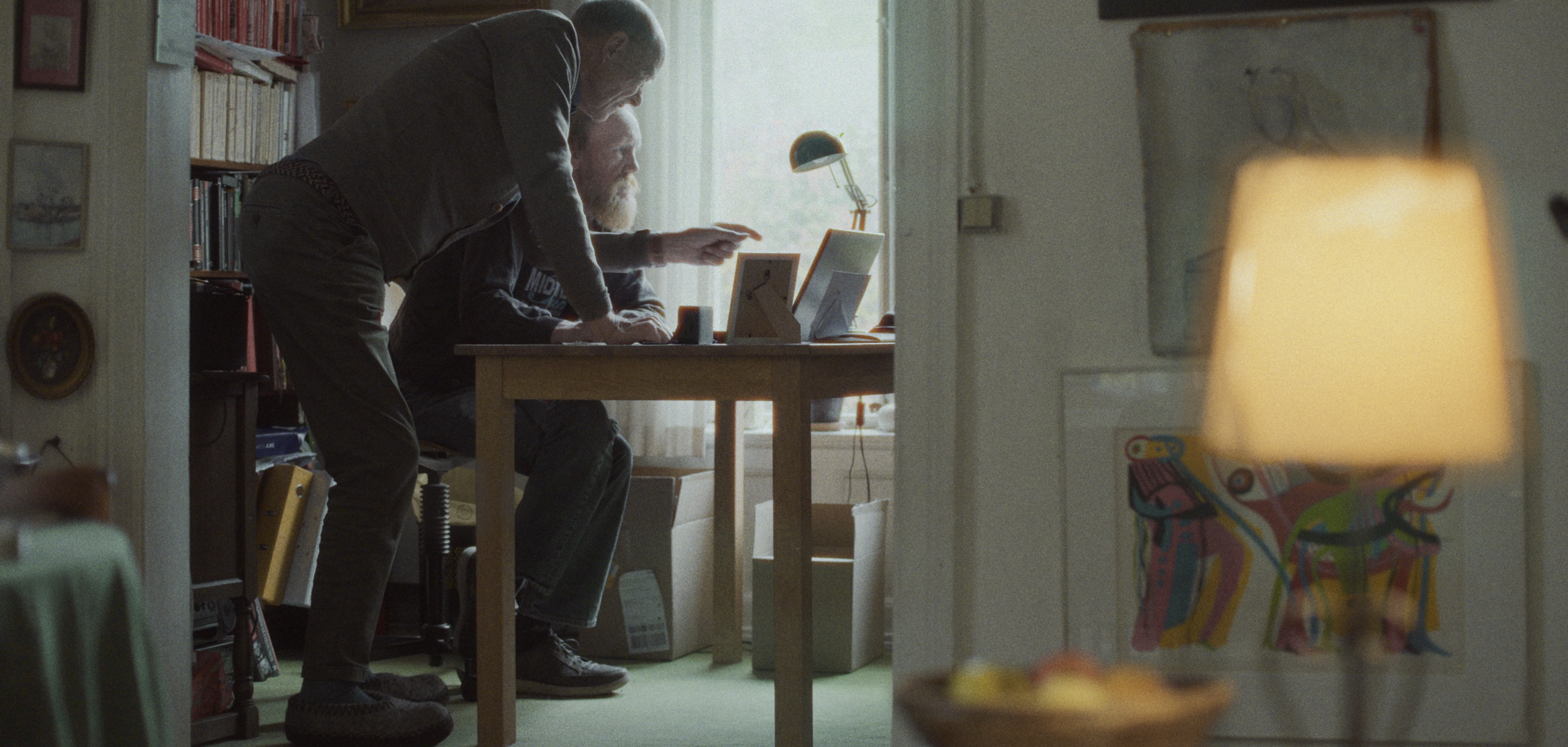 To mænd arbejder sammen ved et bord i et lyst værelse med bøger og billeder på væggen. En lampe er tændt, og der er en skål med frugt i forgrunden | Dokumentarfilm | Retrograd | Esben Persson & Anders Mielcke | Filmproduktion Aarhus | GESTALT