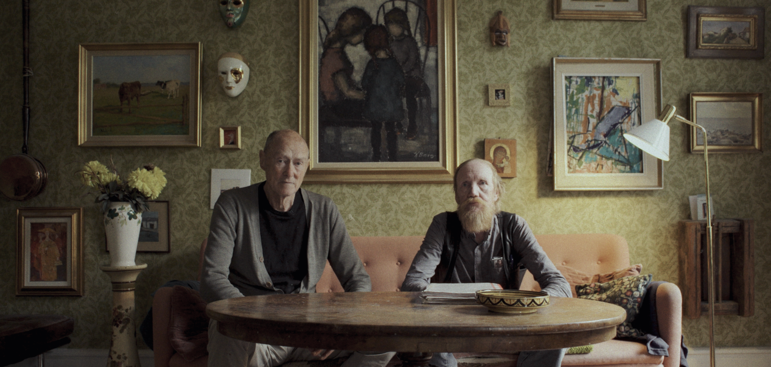 To mænd sidder ved et rundt bord i en stue med vægdekorationer og malerier | Dokumentarfilm | Retrograd | Esben Persson & Anders Mielcke | Filmproduktion Aarhus | GESTALT
