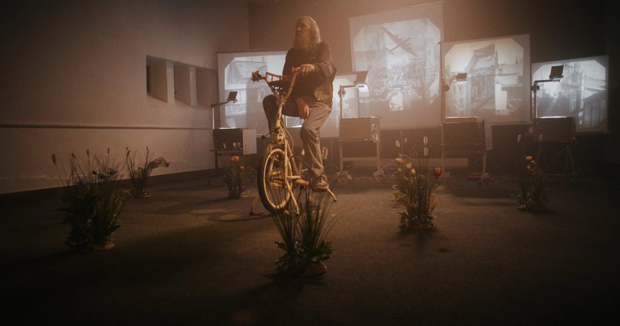 En person med langt hår sidder på en motionscykel,  med blomster omkring, bag skærme, der viser billeder af et boligområde | Dokumentarfilm | Retrograd | Esben Persson & Anders Mielcke | Filmproduktion Aarhus | GESTALT