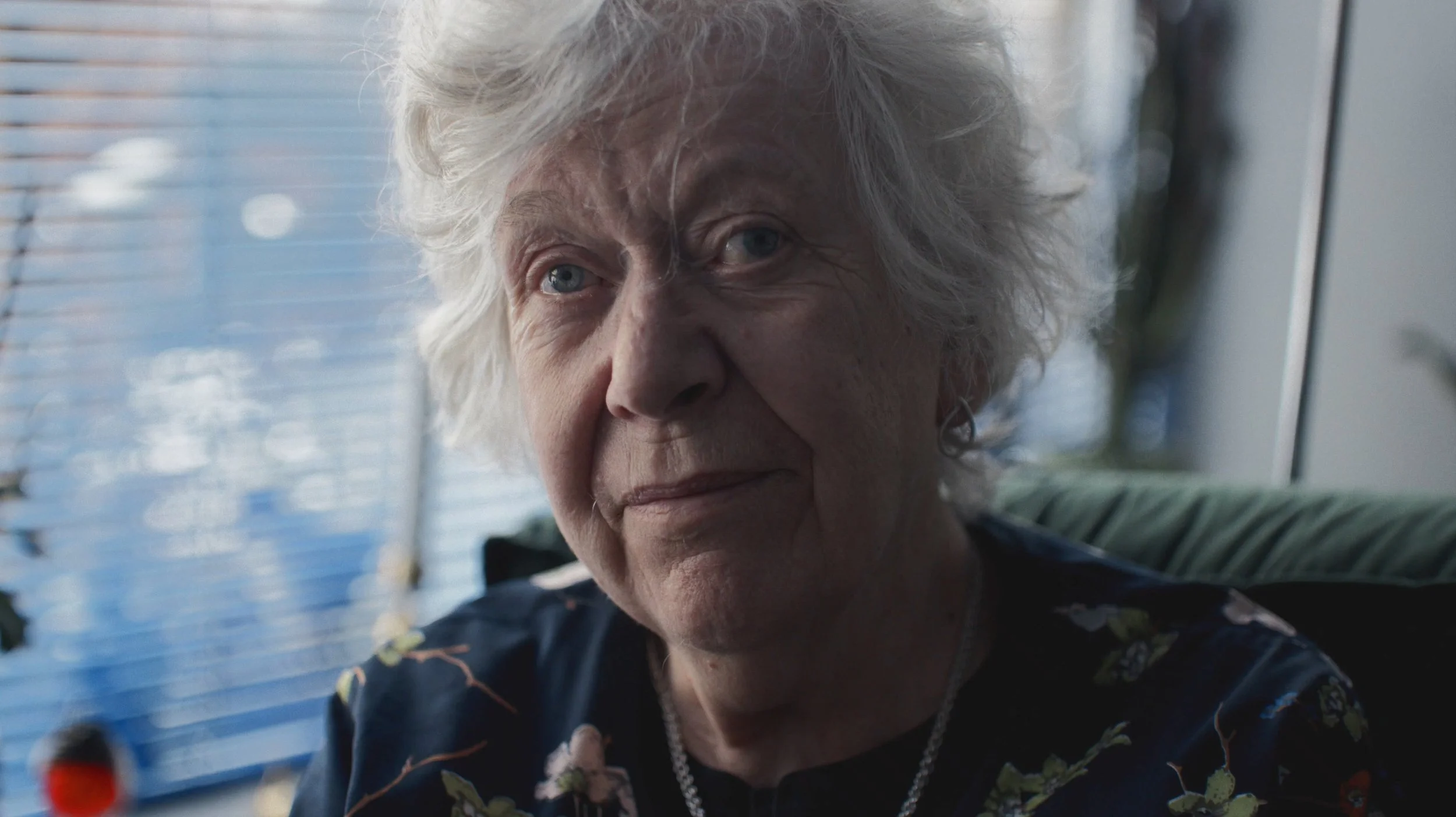 Et nærbillede af en ældre kvinde med gråt, krøllet hår, iført en mørk blomstret top og smykker, siddende foran en vinduesramme | Dokumentarfilm | Stemmer fra sær- og åndssvageforsorgen | Anders Mielcke Grønbæk | GESTALT