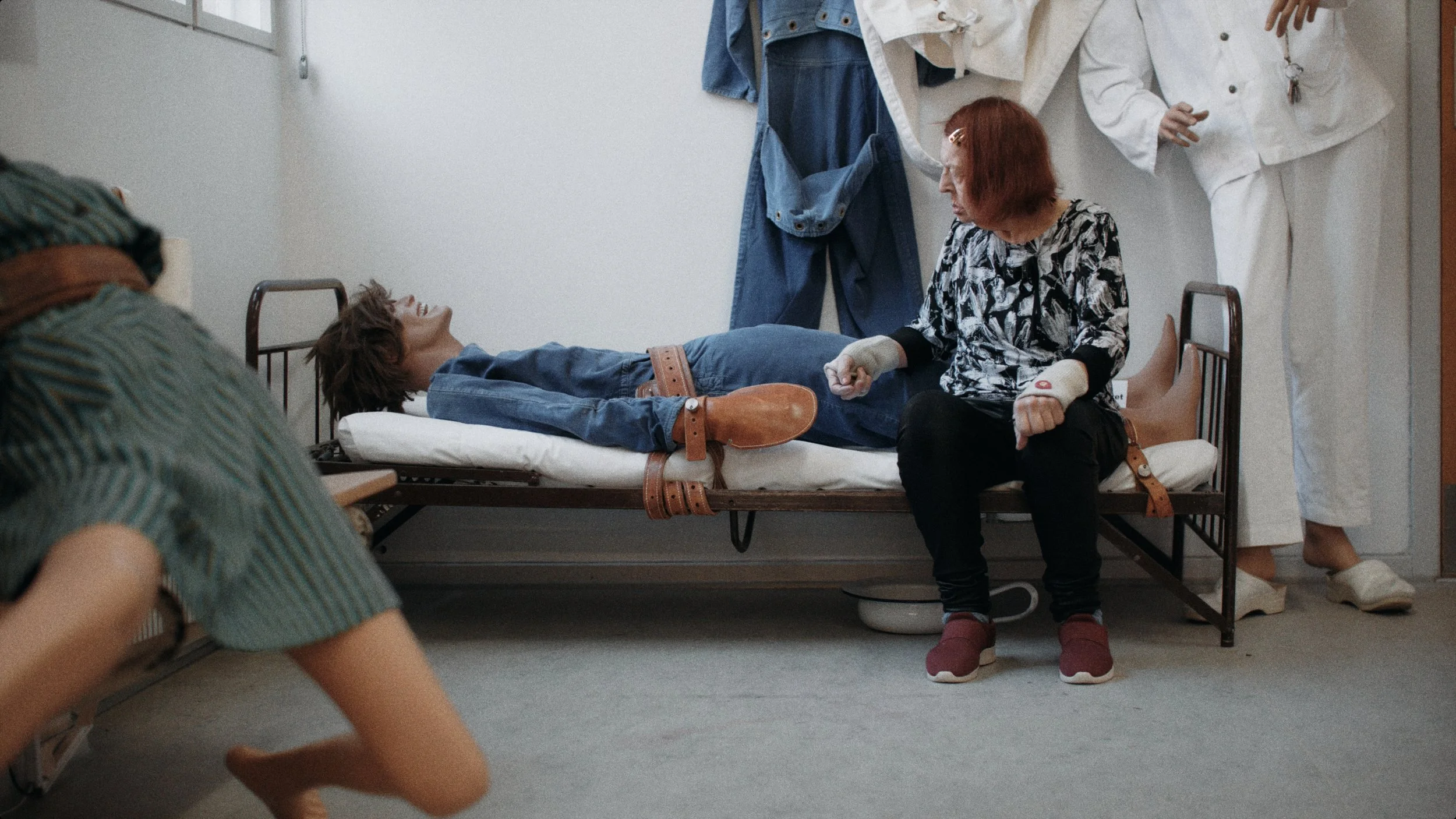 En dukke ligger spændt fast i en hospitalseng. En kvinde sidder ved siden af og holder dukken i hånden i dokumentarisk opstilling | Dokumentarfilm | Stemmer fra sær- og åndssvageforsorgen | Anders Mielcke Grønbæk | GESTALT