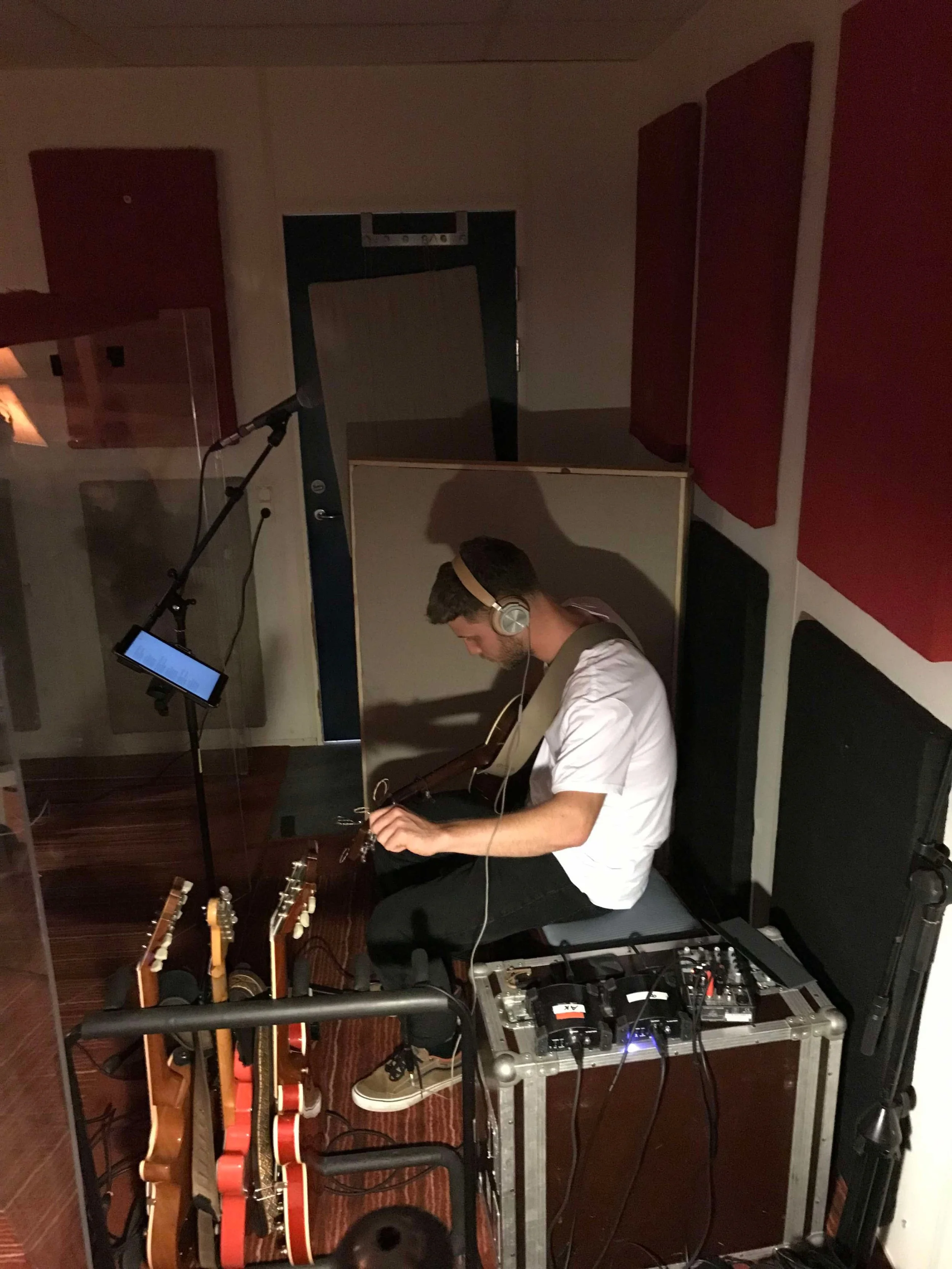 En mand sidder i et lydstudie, spiller guitar og bruger høretelefoner, omgivet af guitarer, lydudstyr og mikrofon | Studieproduktion | Musikkomposition | Filmproduktion Aarhus | GESTALT