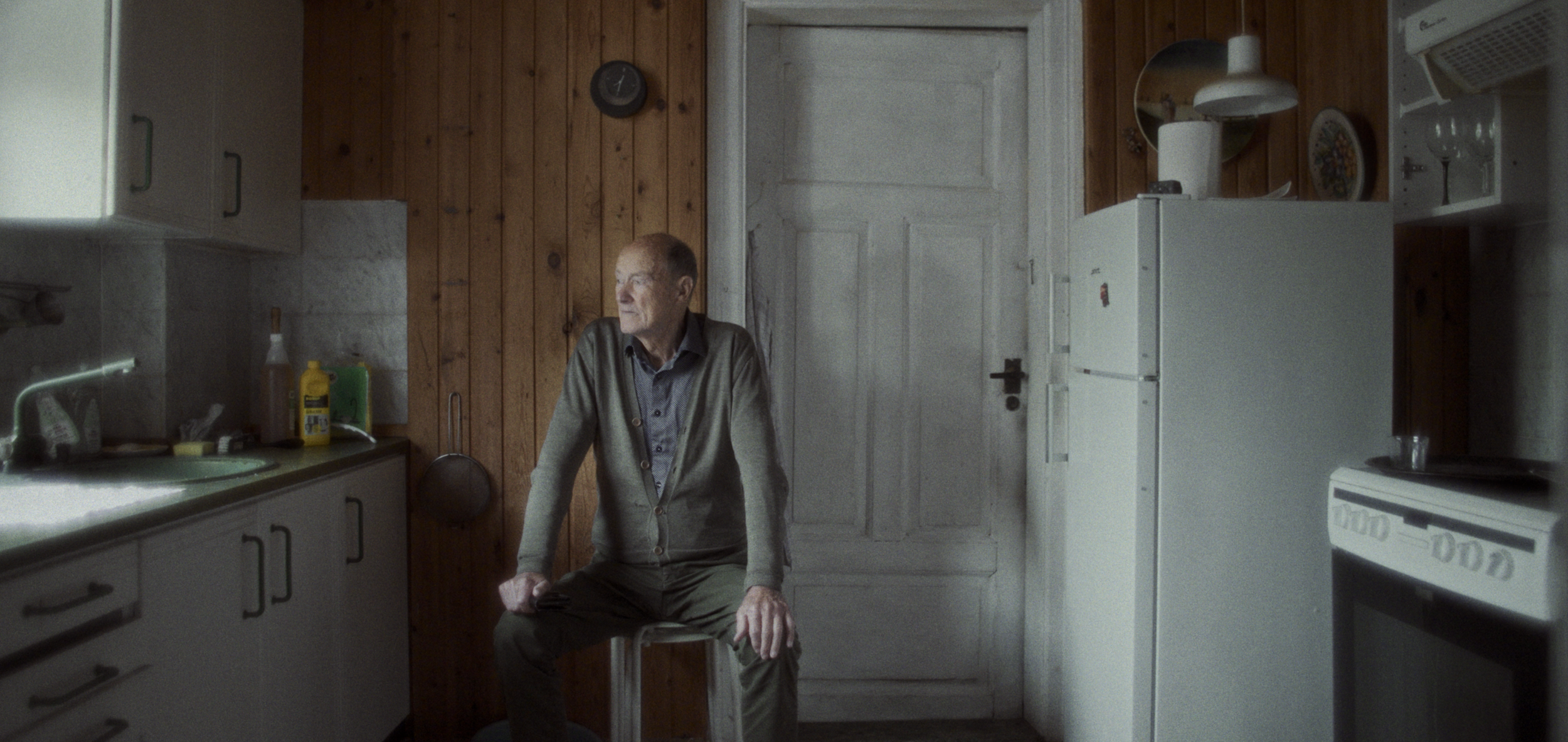 En ældre mand sidder på en stol i et lille, hyggeligt køkken med træpanelvægge | Dokumentarfilm | Retrograd | Esben Persson & Anders Mielcke | Filmproduktion Aarhus | GESTALT