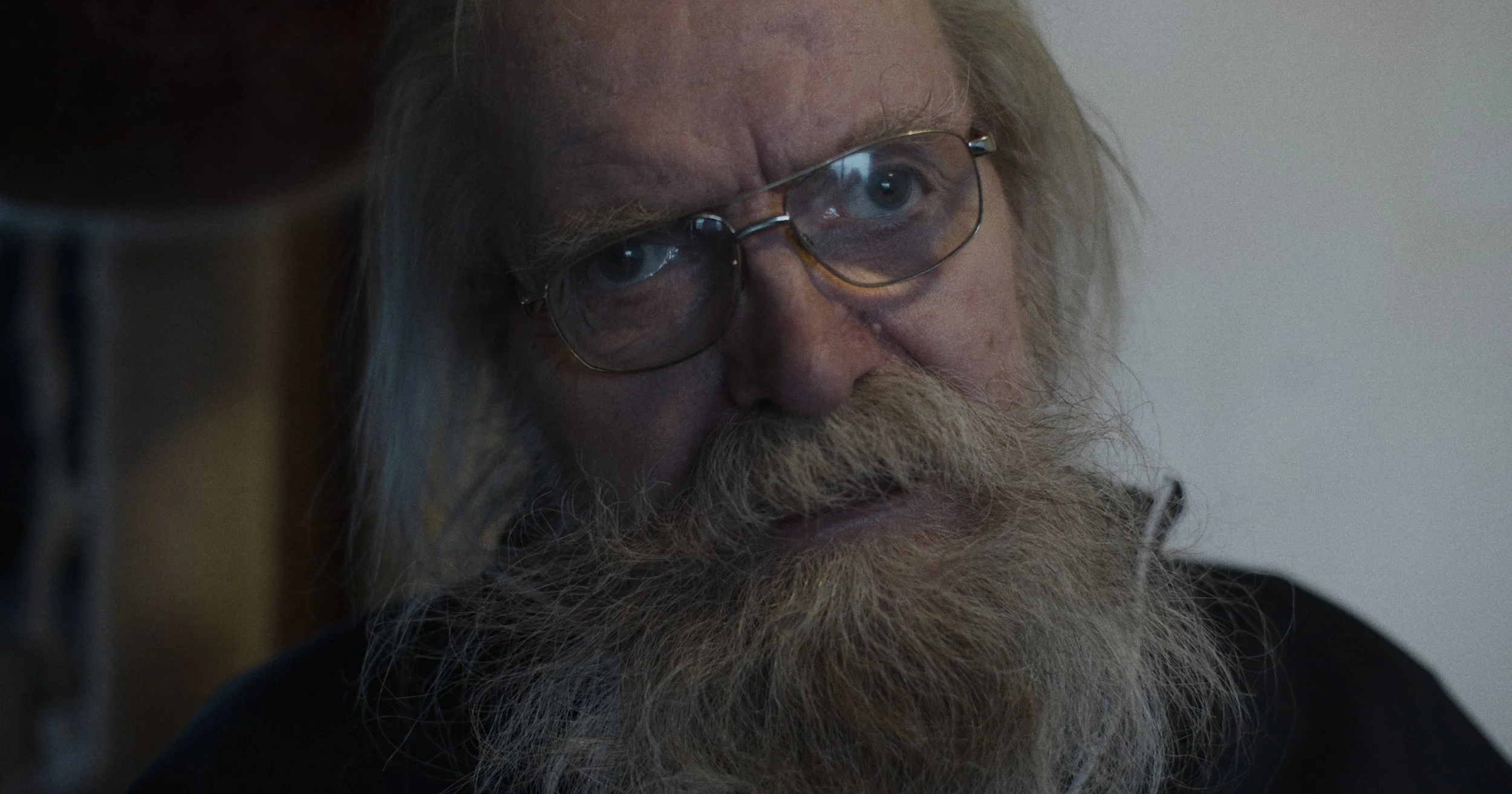 Et nærbillede af en ældre mand med langt, hvidt hår og skæg, der ser tænksom ud, iført briller | Dokumentarfilm | Retrograd | Esben Persson & Anders Mielcke | Filmproduktion Aarhus | GESTALT