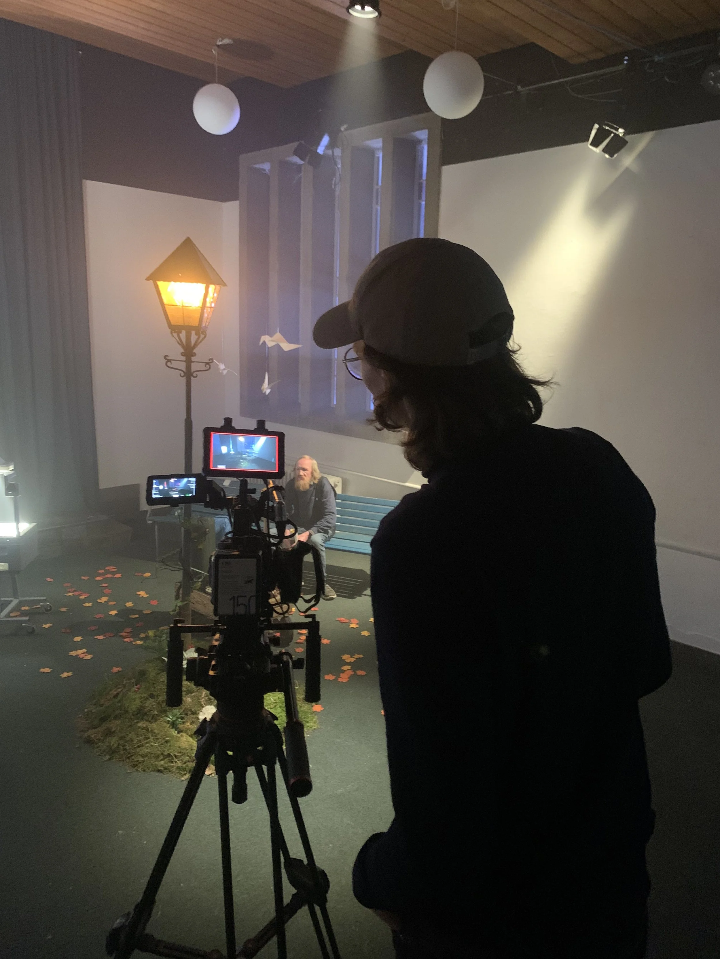 Person, der filmer en scene med en mand med langt skæg og lyse bukser, sidder på en bænk, omgivet af lamper og dekorative elementer, i et indendørs studie | Dokumentarfilm | Retrograd | Esben Persson & Anders Mielcke | Filmproduktion Aarhus | GESTALT