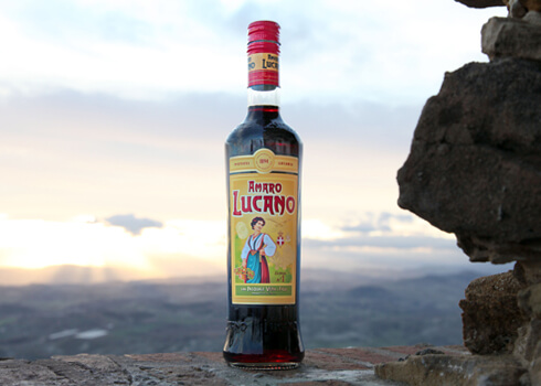 A bottle of Amaro Lucano herbal liqueur on a stone surface with a sunset sky in the background.