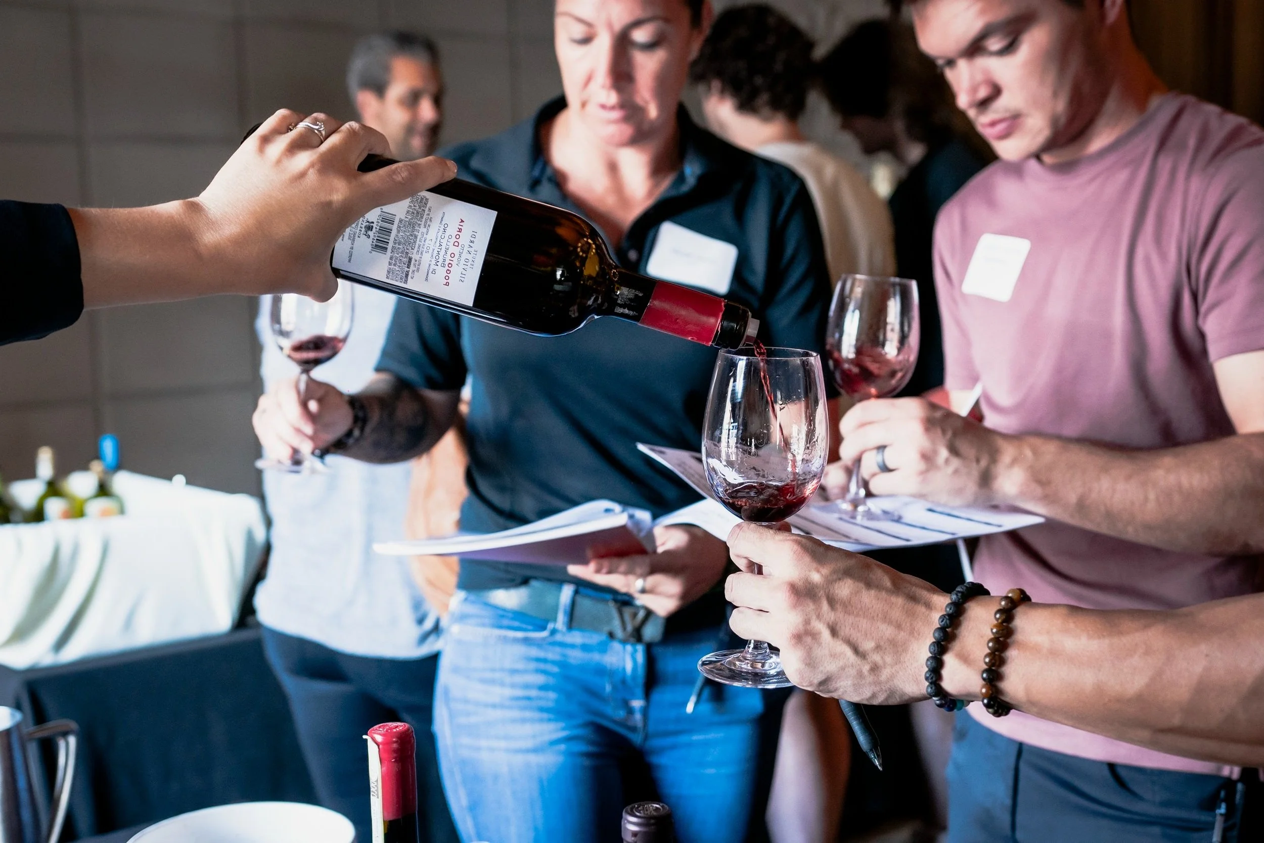 Person pouring red wine into a glass at a tasting event, with other people observing and holding glasses, in an indoor setting.