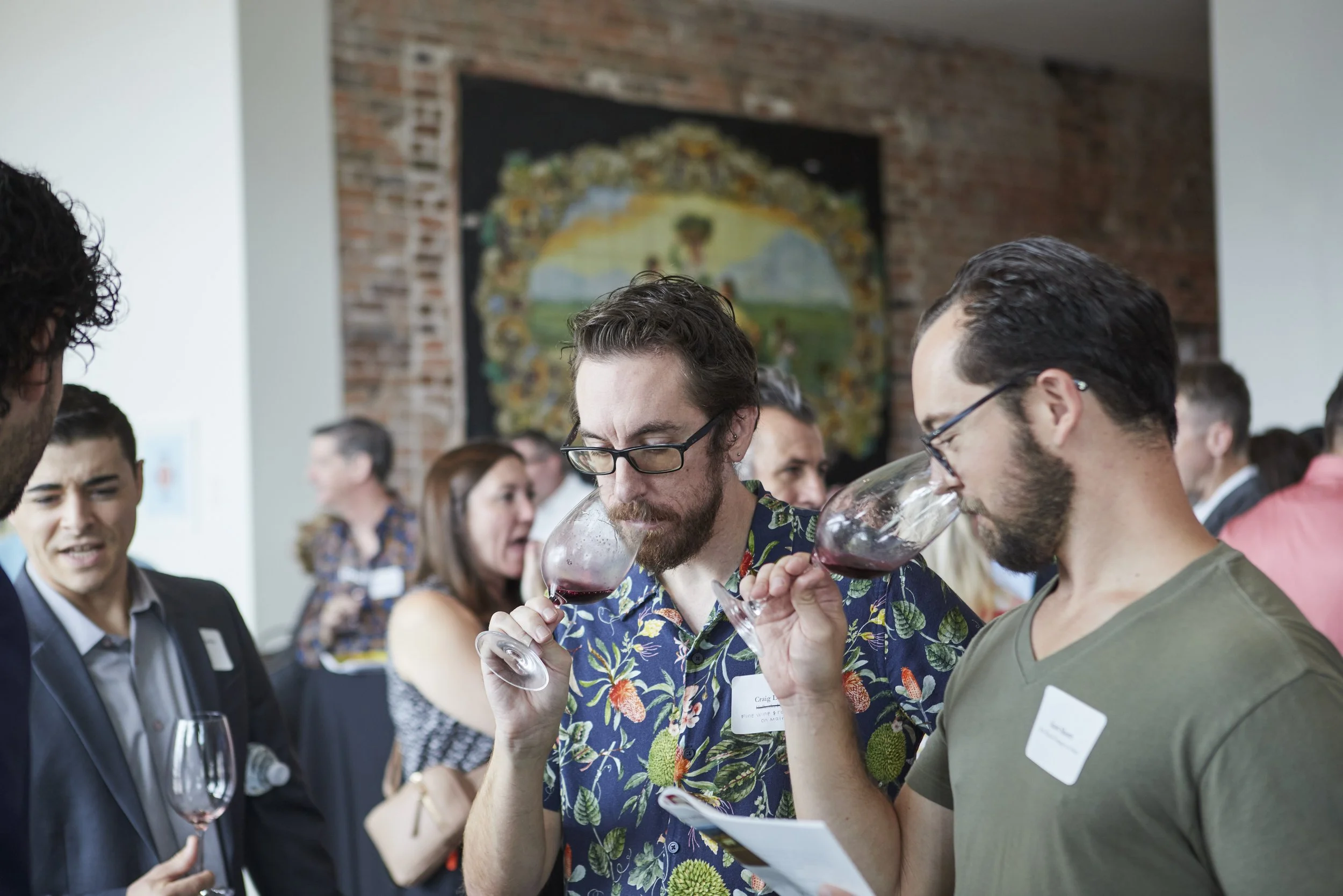 People at a social event holding and drinking wine, engaging in conversation near a brick wall with a colorful painting in the background.