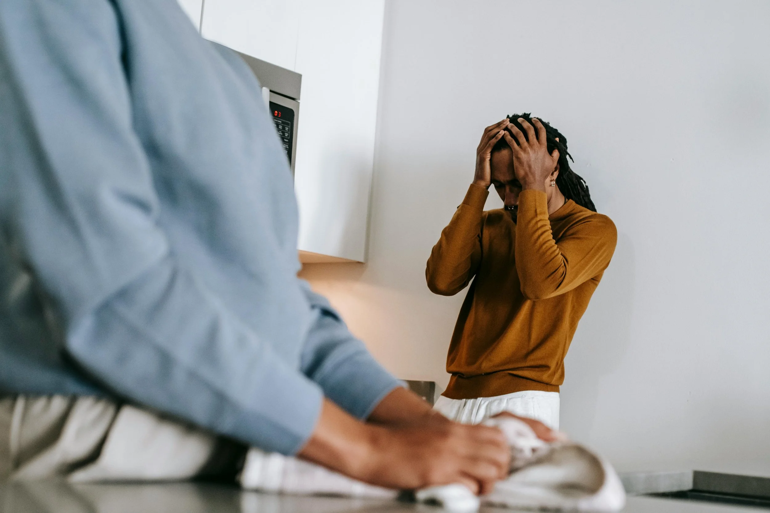 A woman with long braids, wearing an orange sweater and white pants, in distress, holding her head with both hands in a room, while a person in a blue hoodie kneels beside her, handling some cloth or towel on a countertop.