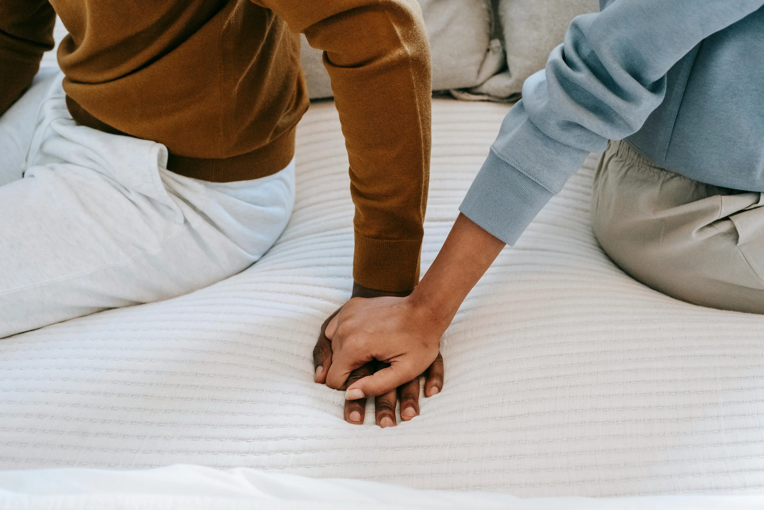 Two people kneeling on a bed holding hands, one wearing a brown long-sleeve shirt and white pants, and the other wearing a blue long-sleeve shirt and beige pants.