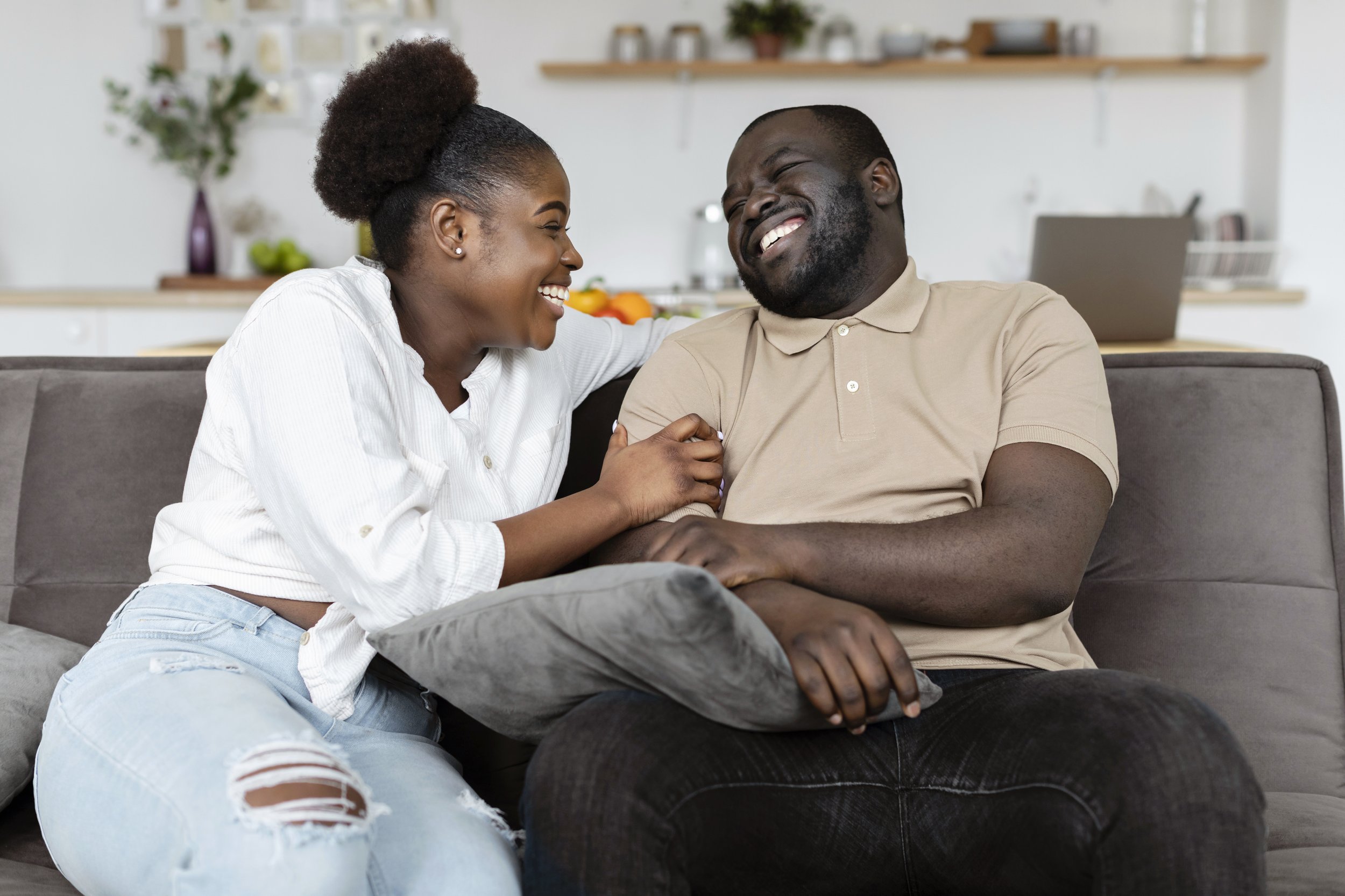 A happy couple sitting on a gray couch in a modern kitchen, laughing and enjoying each other's company.